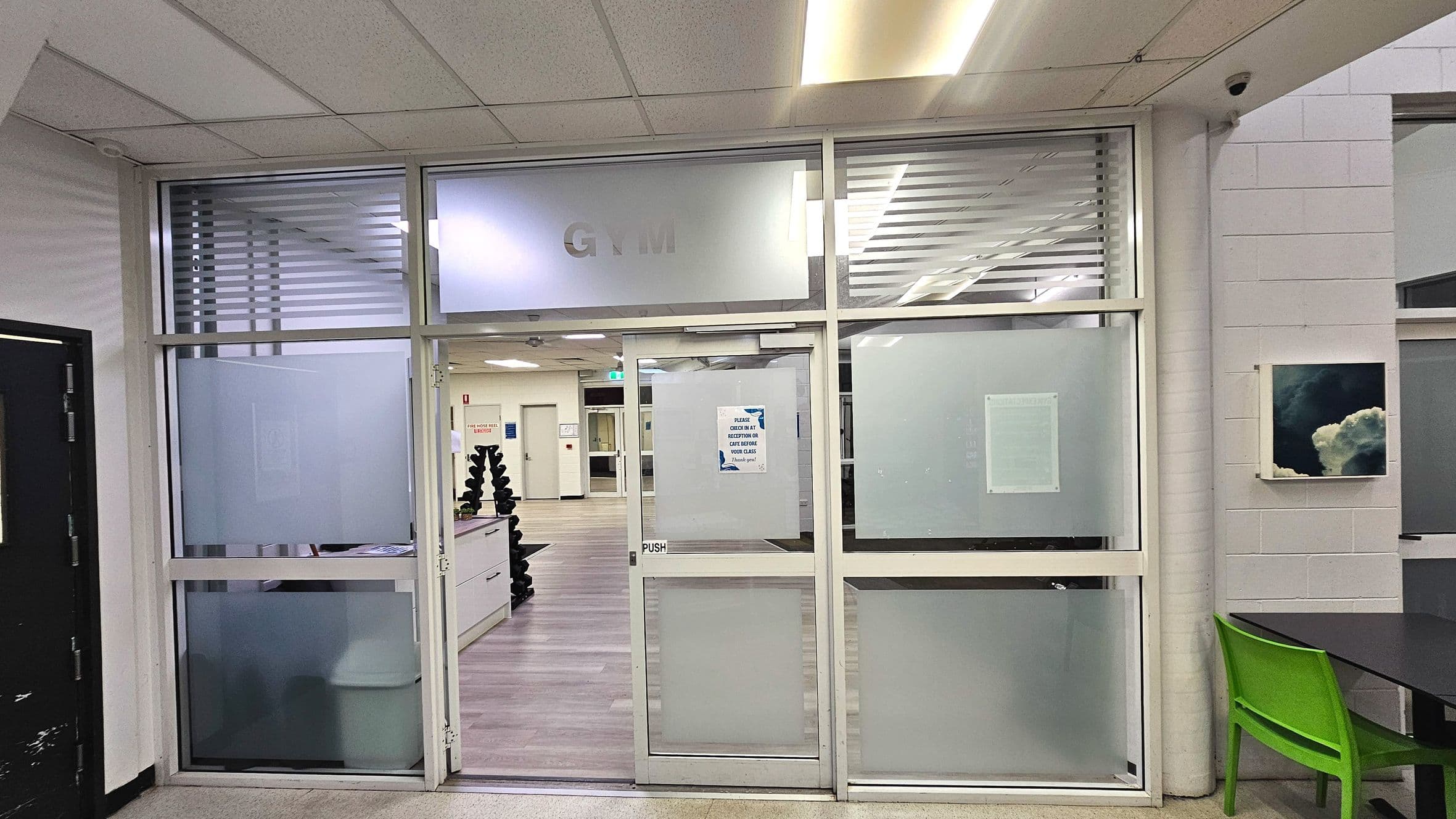 A gym entrance with frosted glass panels and a door marked 'PUSH'. Inside, a wooden floor leads to a reception desk with a stack of black weights visible. The ceiling has fluorescent lighting, and a green chair is placed next to a black table on the right. A small artwork is mounted on the adjacent wall. The space feels enclosed by glass and walls, with a transition from tiled to wooden flooring.
