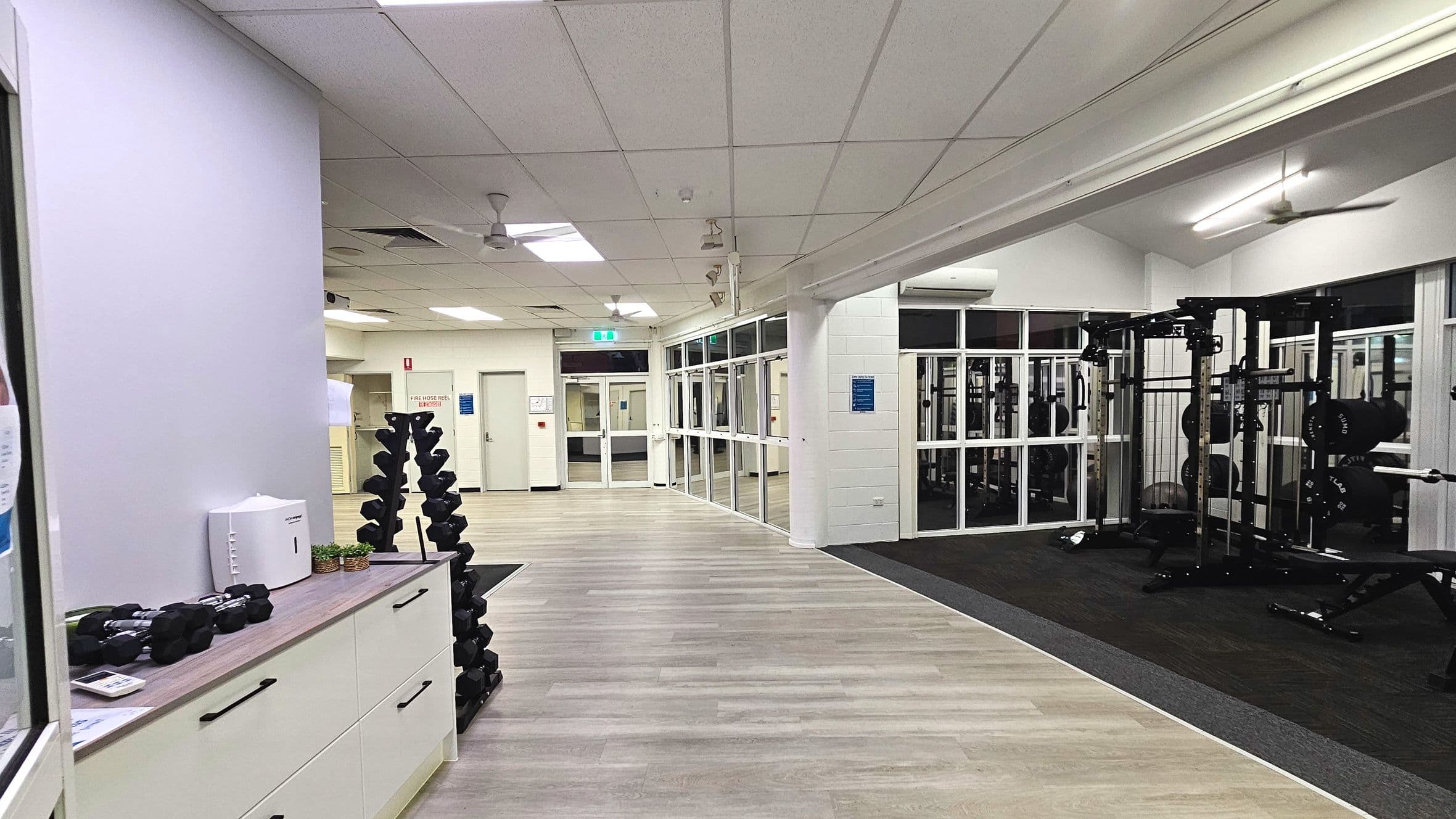 A gym interior with a light wood laminate floor transitioning to a dark carpeted area. On the left, a white cabinet with black handles holds small dumbbells and a white device. A vertical rack of dumbbells stands nearby. The right side features weightlifting equipment. The space is enclosed by white walls and large glass panels. Ceiling fans and fluorescent lights are visible above.