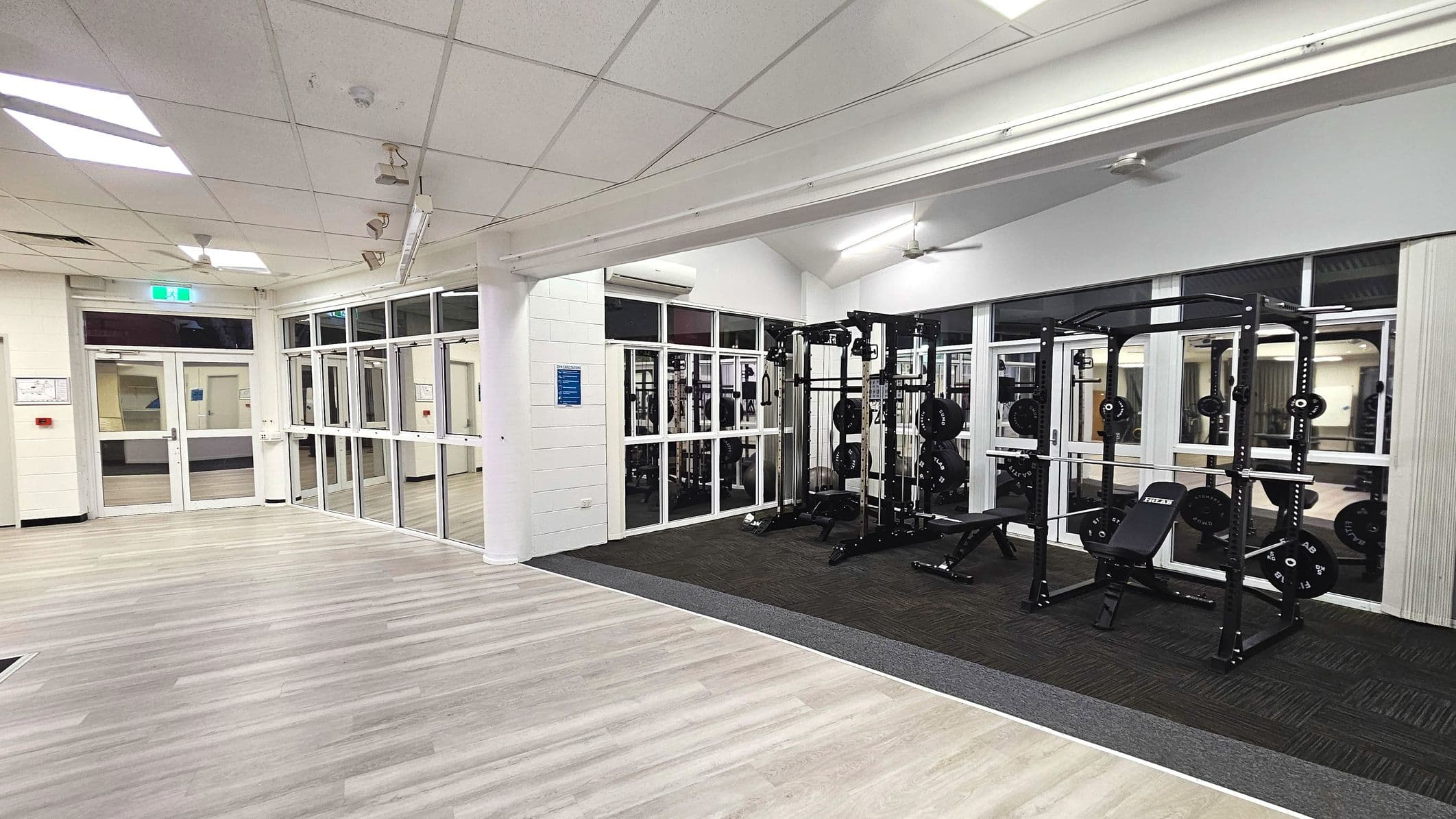 A gym area with weightlifting equipment is visible, featuring two squat racks with weights and benches on a dark carpeted section. Adjacent is a spacious area with light wood flooring. The space is enclosed by large glass windows and doors, providing a view into another room. The ceiling is fitted with fluorescent lights and ceiling fans. An emergency exit sign is above a door in the background.