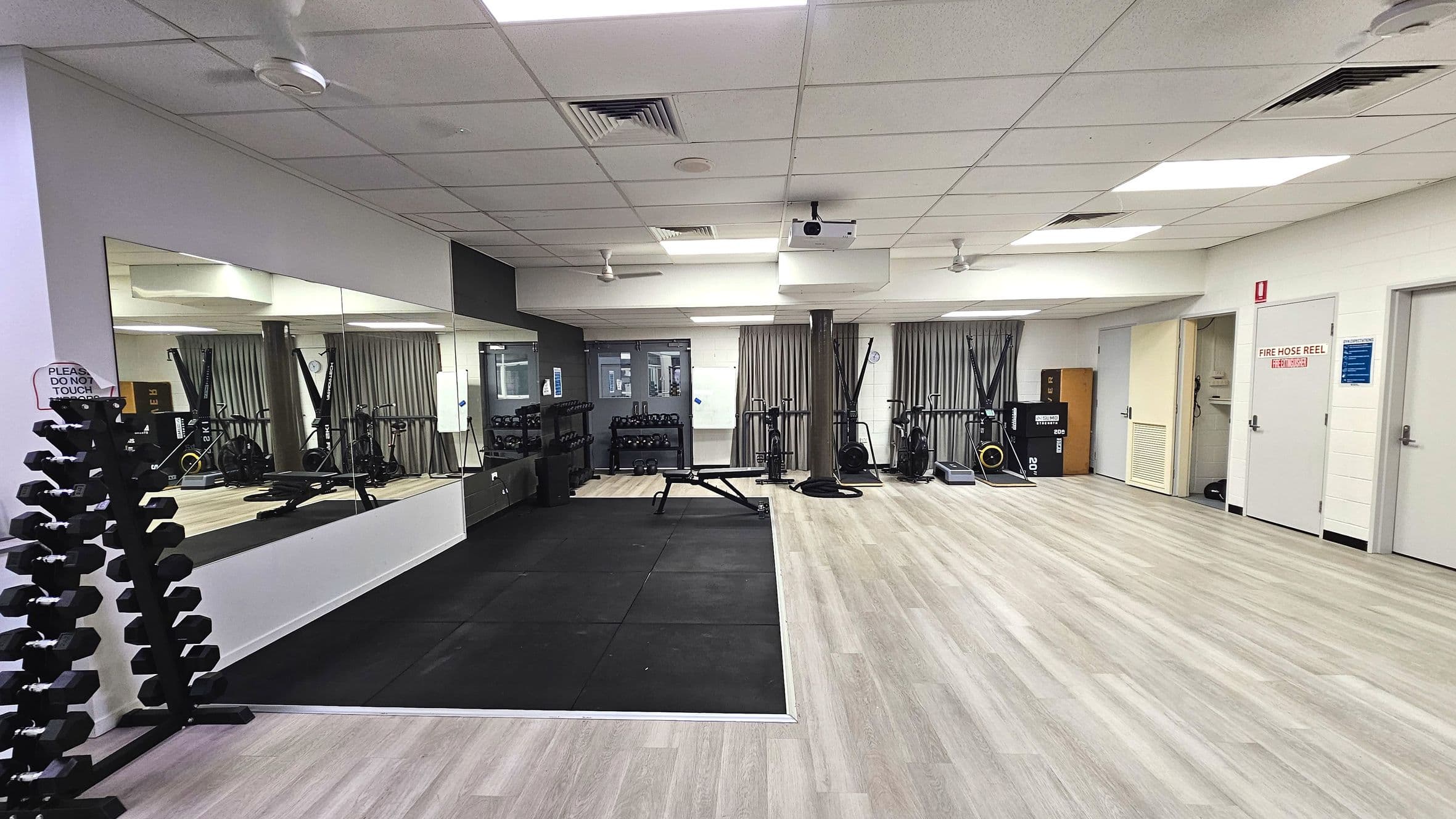 A gym room with a large wall mirror reflecting exercise equipment. On the left, a rack of dumbbells stands on a light wood floor. Adjacent is a black rubber mat area with a bench. The right side features exercise bikes and a rowing machine. The ceiling has recessed lighting and ventilation panels. Doors with signage, including a fire hose reel, line the right wall. Curtains cover windows at the back.