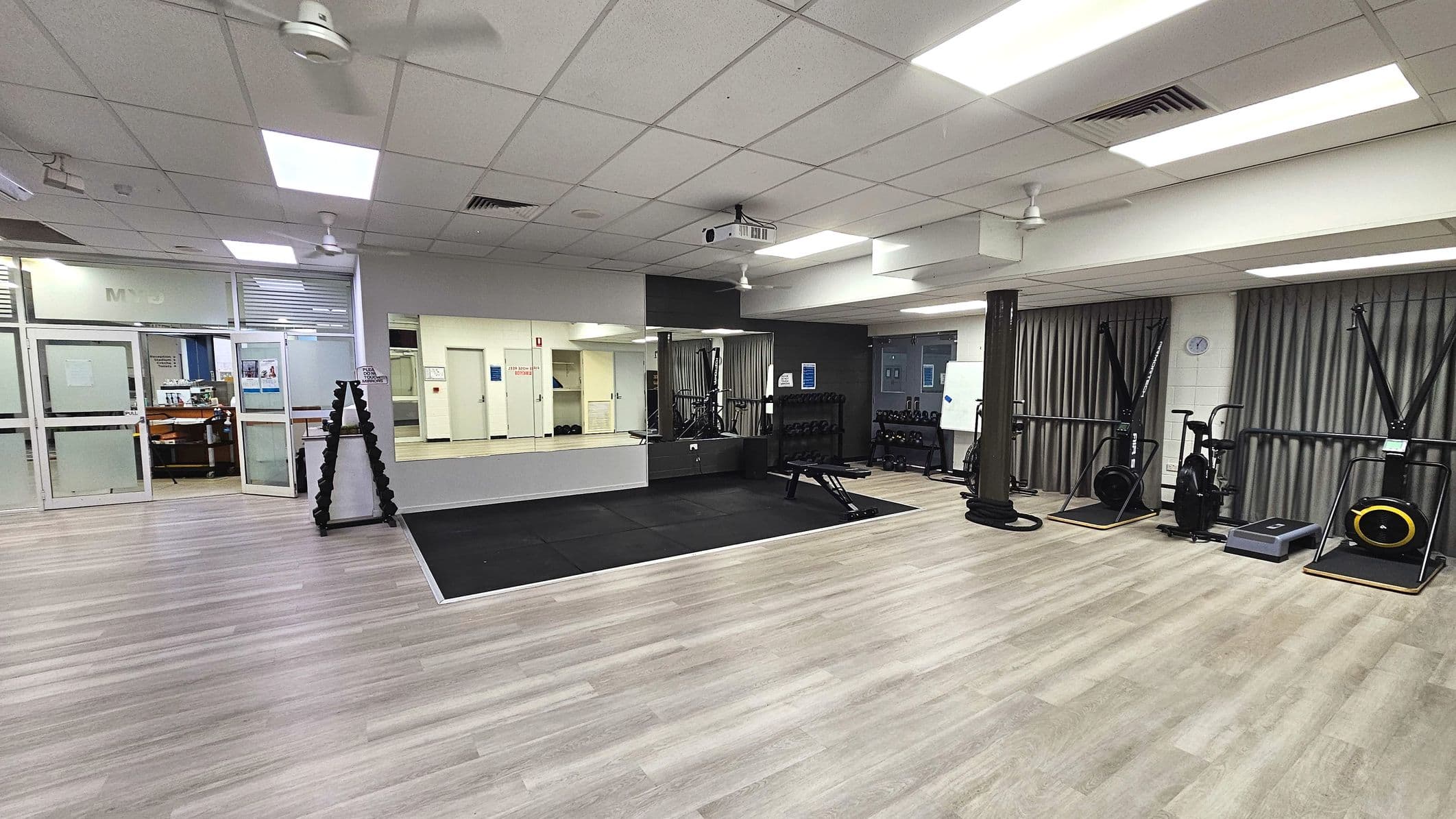 A spacious gym room with a light wood laminate floor and a section of black rubber matting. The room features various exercise equipment including stationary bikes and a step platform. A large mirror covers one wall, reflecting the equipment. Ceiling fans and fluorescent lights are visible above. A glass door with frosted panels leads to another area. A rack of dumbbells is positioned near the mirror.