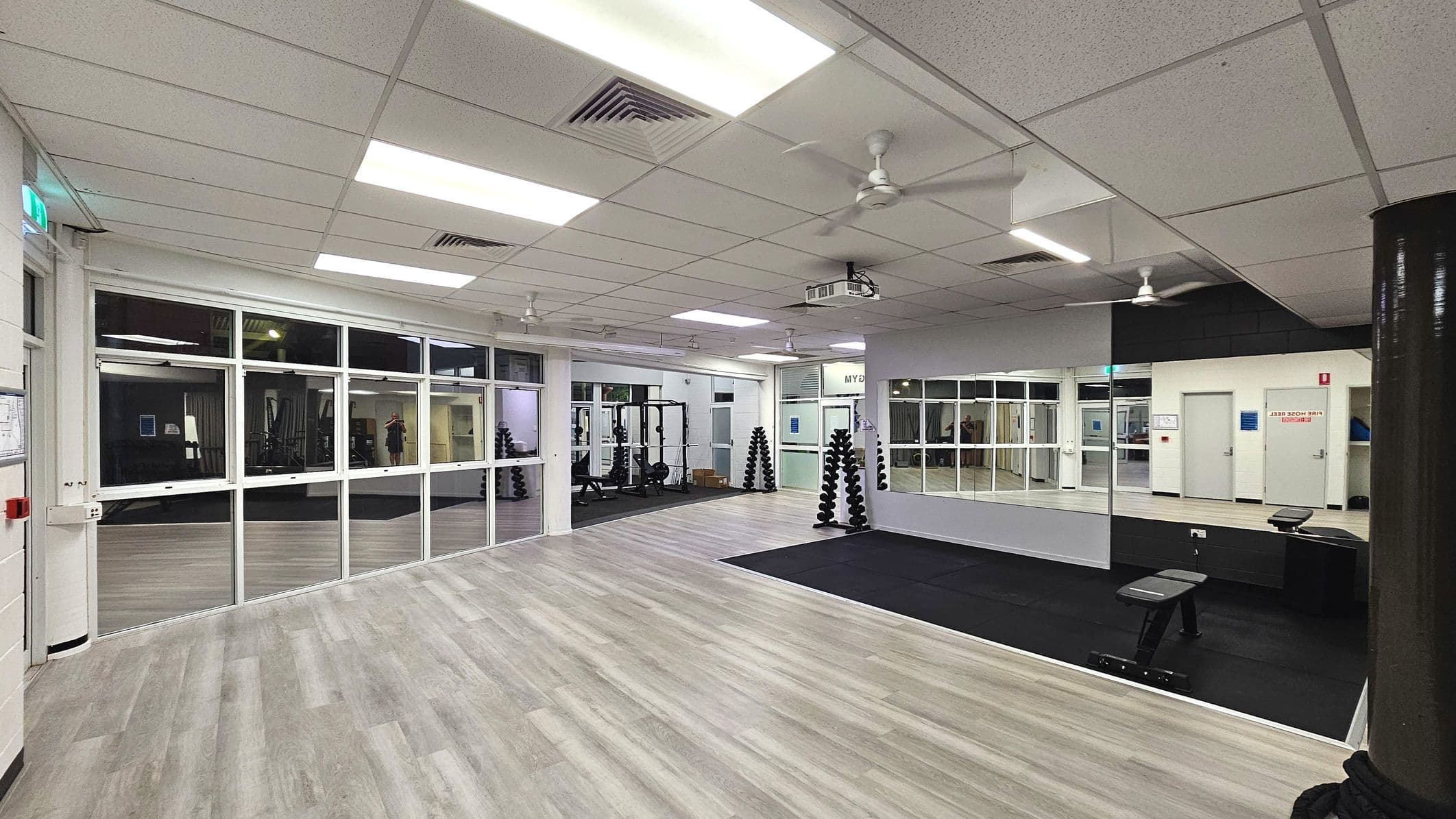 A spacious gym area with light wood laminate flooring and a section of black matting. The room features large glass windows and mirrors along the walls, enhancing the sense of openness. Several racks of dumbbells are neatly arranged, and a weight bench is positioned on the matting. The ceiling is fitted with fluorescent lights and ceiling fans. Doors and safety signage are visible on the walls, indicating exits and fire safety information.