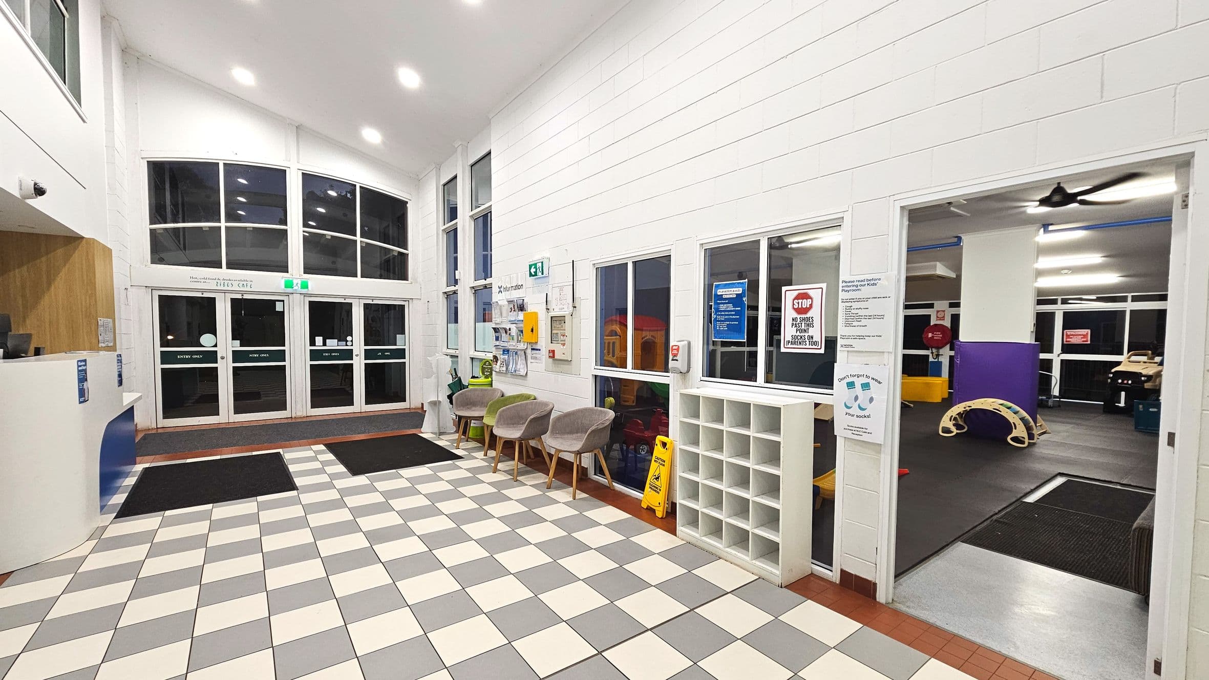 A spacious indoor area with a high ceiling and large windows. The floor is tiled in a checkerboard pattern of grey and cream. On the left, a reception desk is visible. To the right, a row of beige chairs is placed against the wall, next to a shoe cubby. A doorway leads to a play area with a rubber floor and children's equipment. Double glass doors at the back serve as the main entrance, flanked by informational signage.