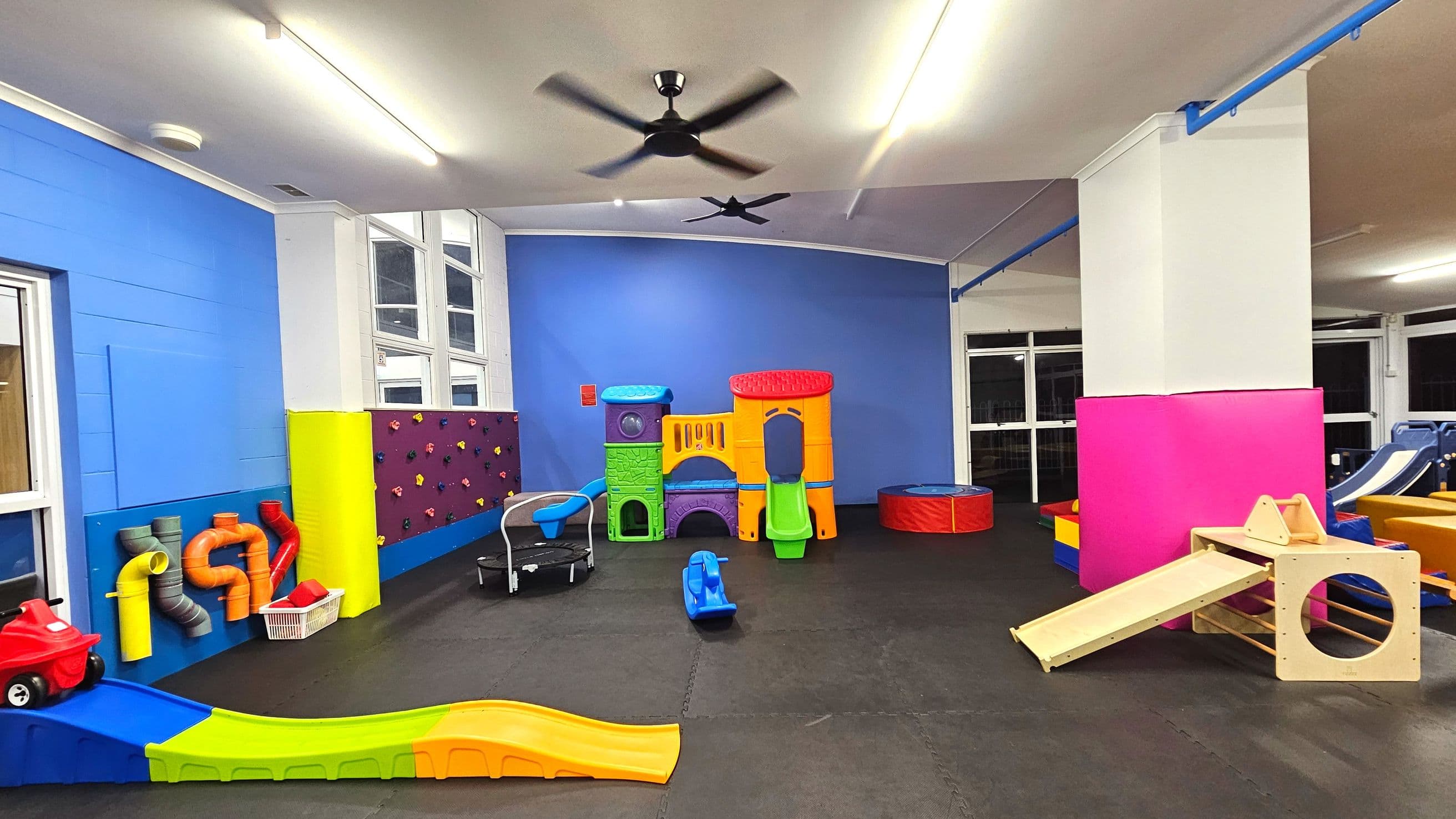 A vibrant indoor play area with colourful equipment. A large plastic play structure with slides and tunnels is centred against a blue wall. To the left, a climbing wall with multicoloured grips and a small trampoline are visible. A wooden slide and cube structure are on the right. The floor is covered with black rubber mats, and the ceiling has fluorescent lights and a fan. Large windows line the walls, providing natural light.