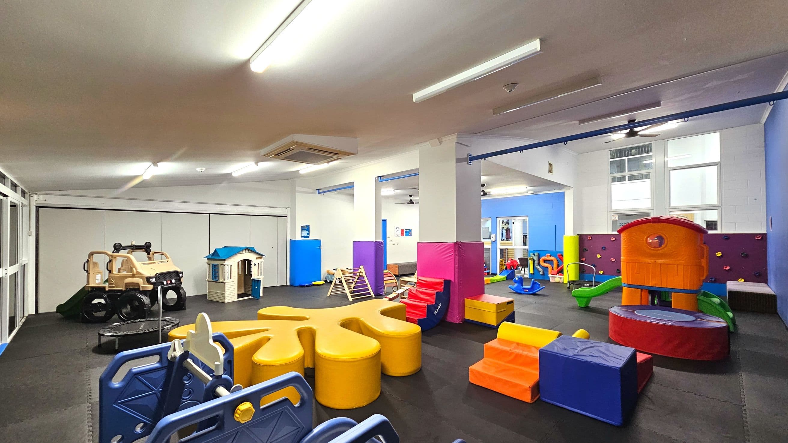 A spacious indoor play area with various colourful play structures. On the left, a beige toy car and a small playhouse are visible. The centre features a yellow star-shaped seating area and blue and red padded steps. To the right, an orange play structure with a slide is set against a wall with climbing holds. The floor is covered with black foam mats, and the room is well-lit with fluorescent lights and large windows.