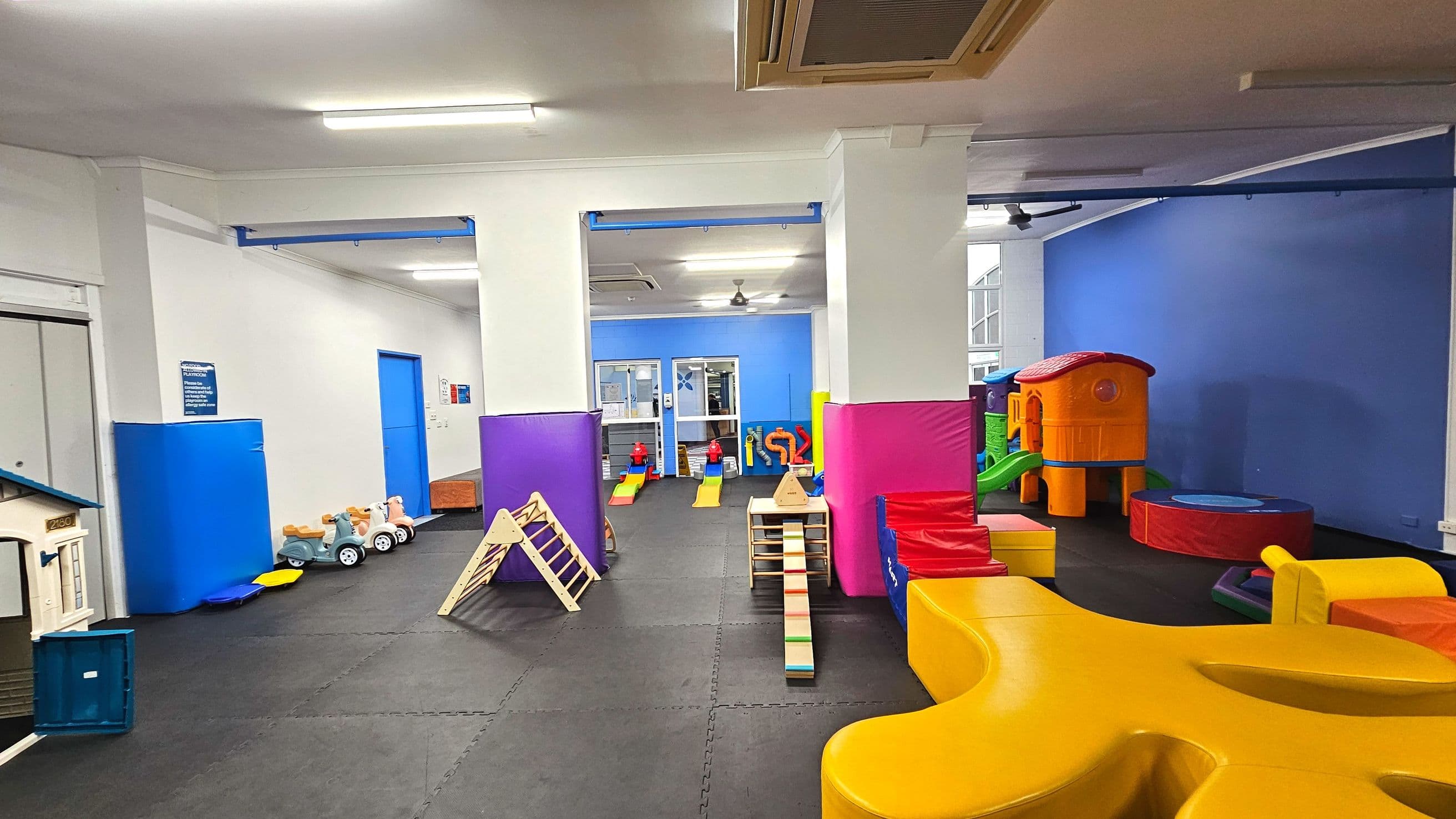 A spacious indoor play area with colourful padded flooring and walls. The room features various play structures including a small plastic house, climbing frames, slides, and soft play equipment in bright colours like yellow, red, and purple. The walls are painted blue and white, and there are large pillars wrapped in protective padding. Overhead, fluorescent lights illuminate the space, and a ceiling fan is visible.