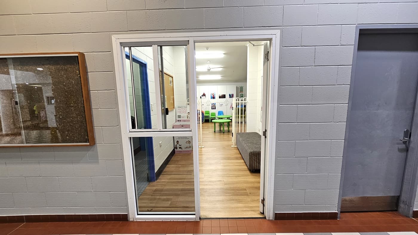 A hallway with light grey brick walls features a glass door leading into a room. The door has a white frame and is partially open, revealing a space with wooden flooring and a green table surrounded by colourful chairs. A bulletin board is mounted on the left wall, and a closed grey door is on the right. The room beyond the door is well-lit with fluorescent lights and decorated with wall posters.