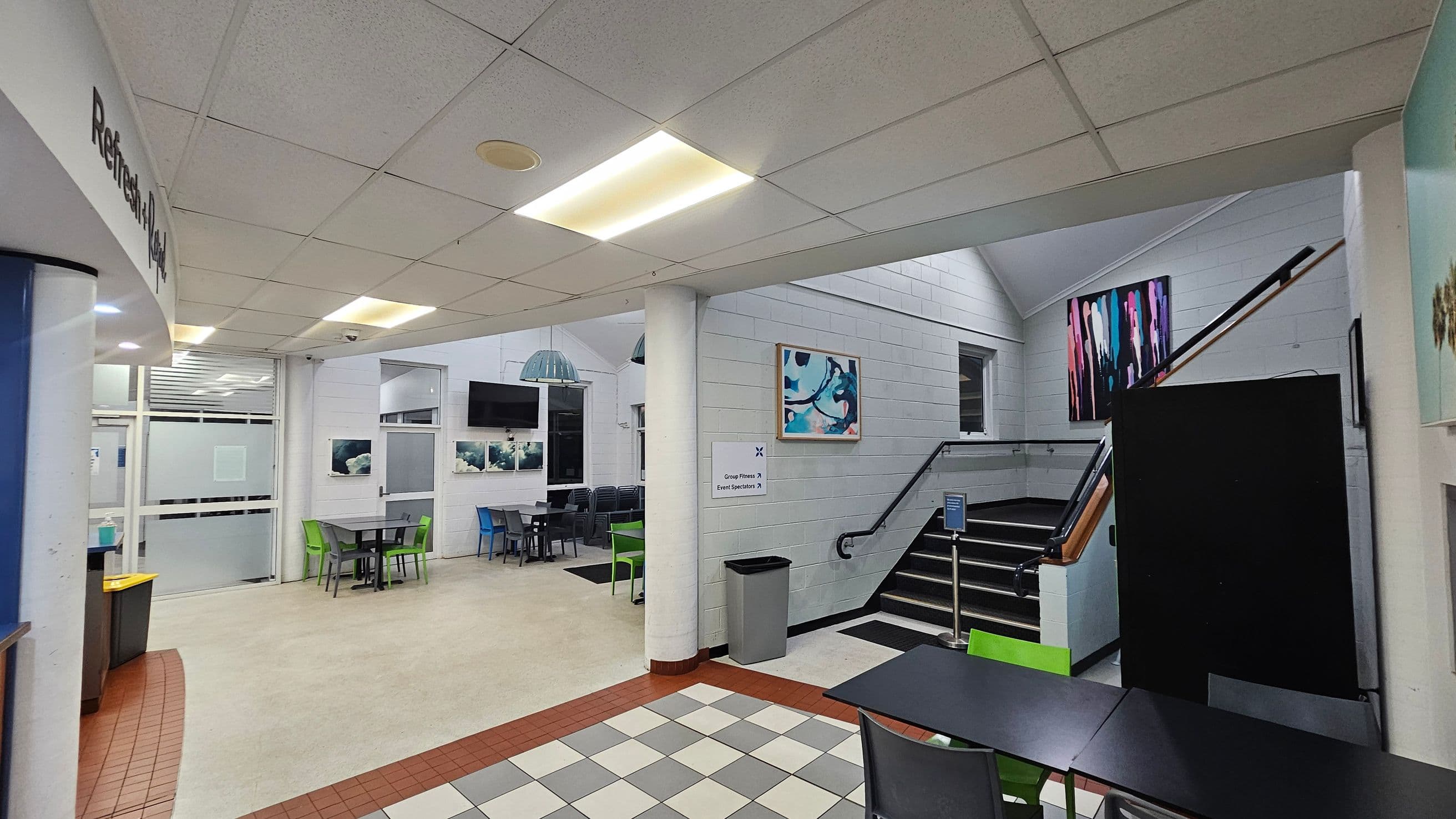 The image shows an indoor space with a tiled floor transitioning to a carpeted area. On the left, a curved wall with 'Refresh' signage leads to a seating area with tables and chairs in various colours. A staircase with a black railing ascends on the right, adorned with colourful abstract art. The walls are white brick, and the ceiling is lined with fluorescent lights. A sign indicates directions to 'Group Fitness' and 'Event Spectator'.