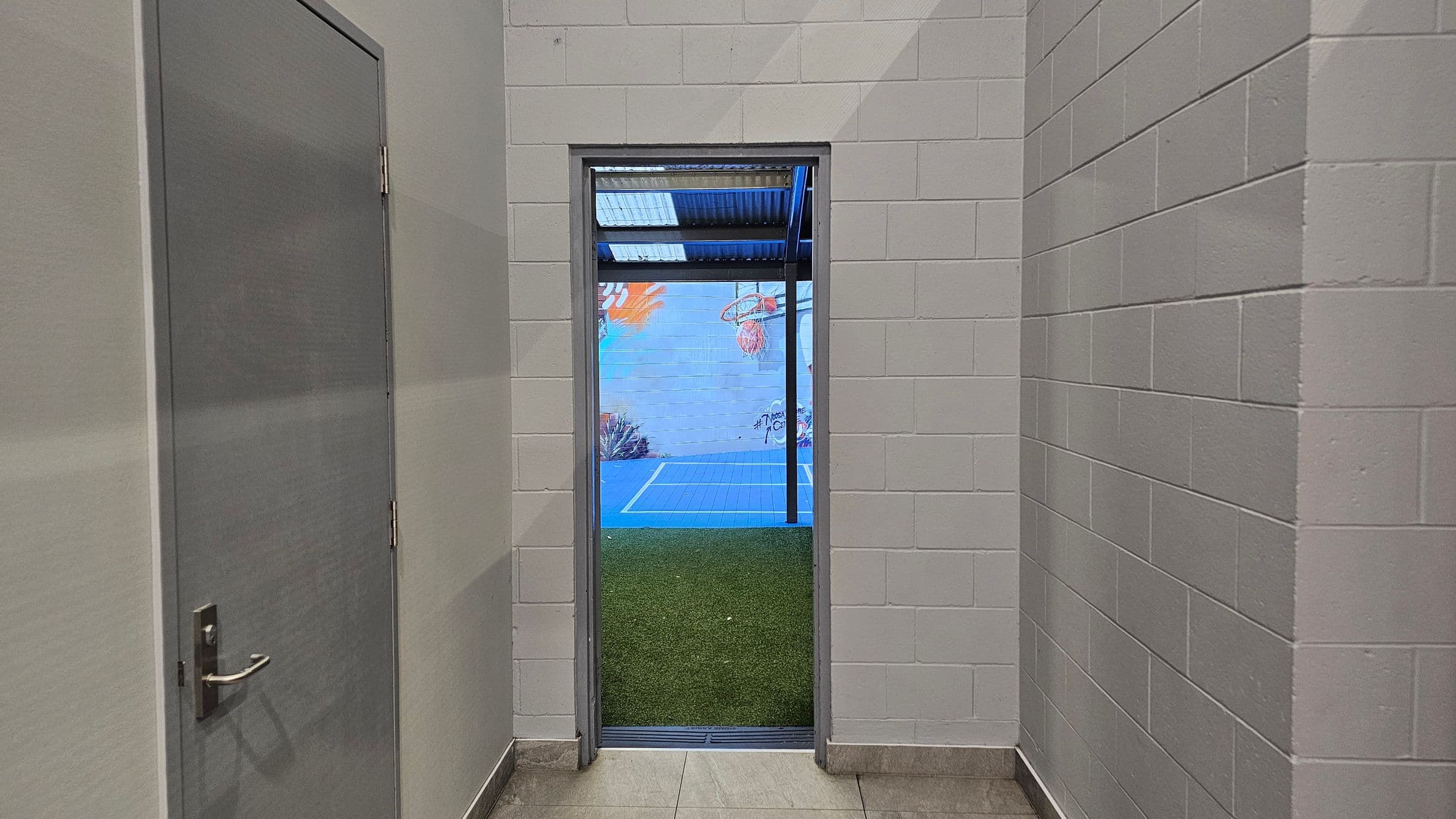 A narrow corridor with light grey brick walls leads to an open doorway. To the left, a closed grey door with a metal handle is visible. Through the doorway, a small outdoor area with artificial grass and a blue sports court is seen. A colourful mural decorates the far wall, featuring abstract shapes and a basketball hoop. The ceiling above the doorway is covered with transparent panels allowing light through.