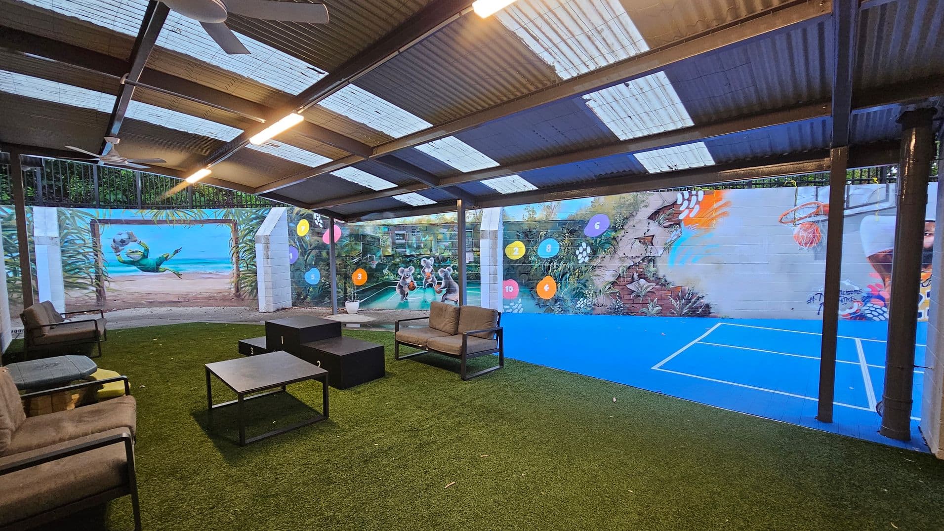 A covered outdoor area with a corrugated metal roof and transparent panels allowing light in. The floor is a mix of artificial grass and a blue sports court. On the left, there are cushioned chairs and tables on the grass. The right wall features vibrant murals of animals and numbers, with a basketball hoop above the blue court. Ceiling fans are mounted under the roof, and the space is open on one side.