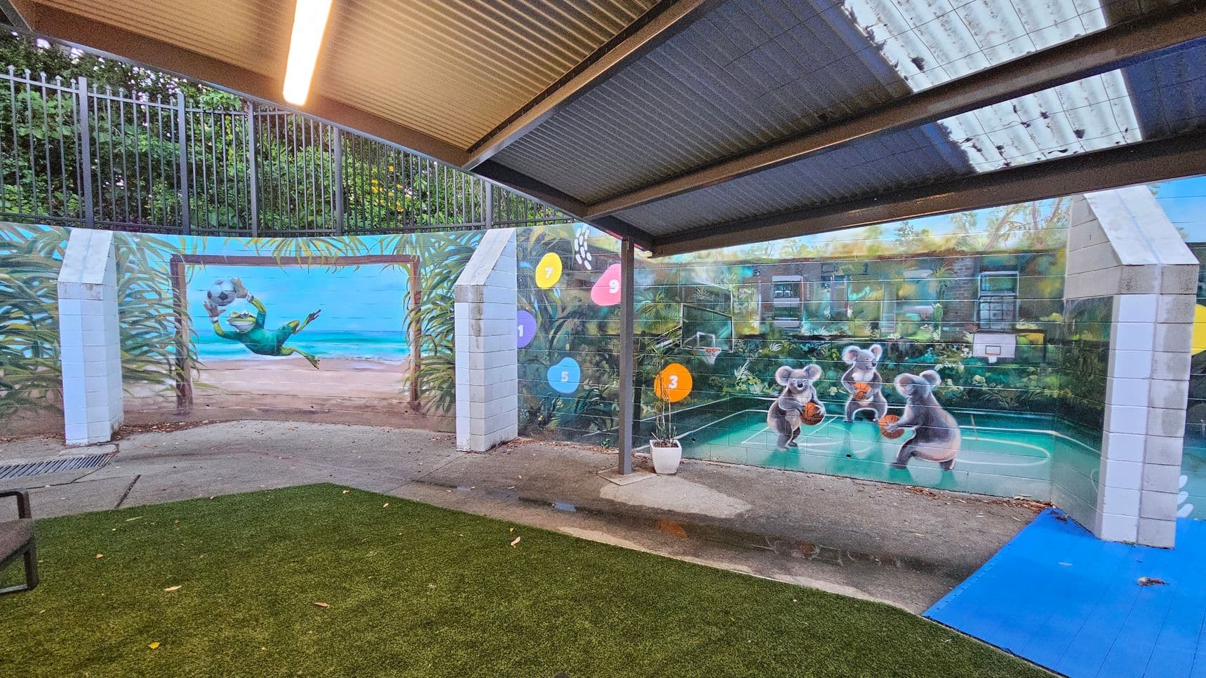 An outdoor area with a corrugated metal roof and a mural on the walls. The mural features a beach scene with a diving goalkeeper and koalas playing basketball on a court. The ground is a mix of artificial grass and concrete, with a blue mat on one side. A metal fence runs along the top of the wall. Coloured circles with numbers are painted on the wall. A chair is partially visible on the left.