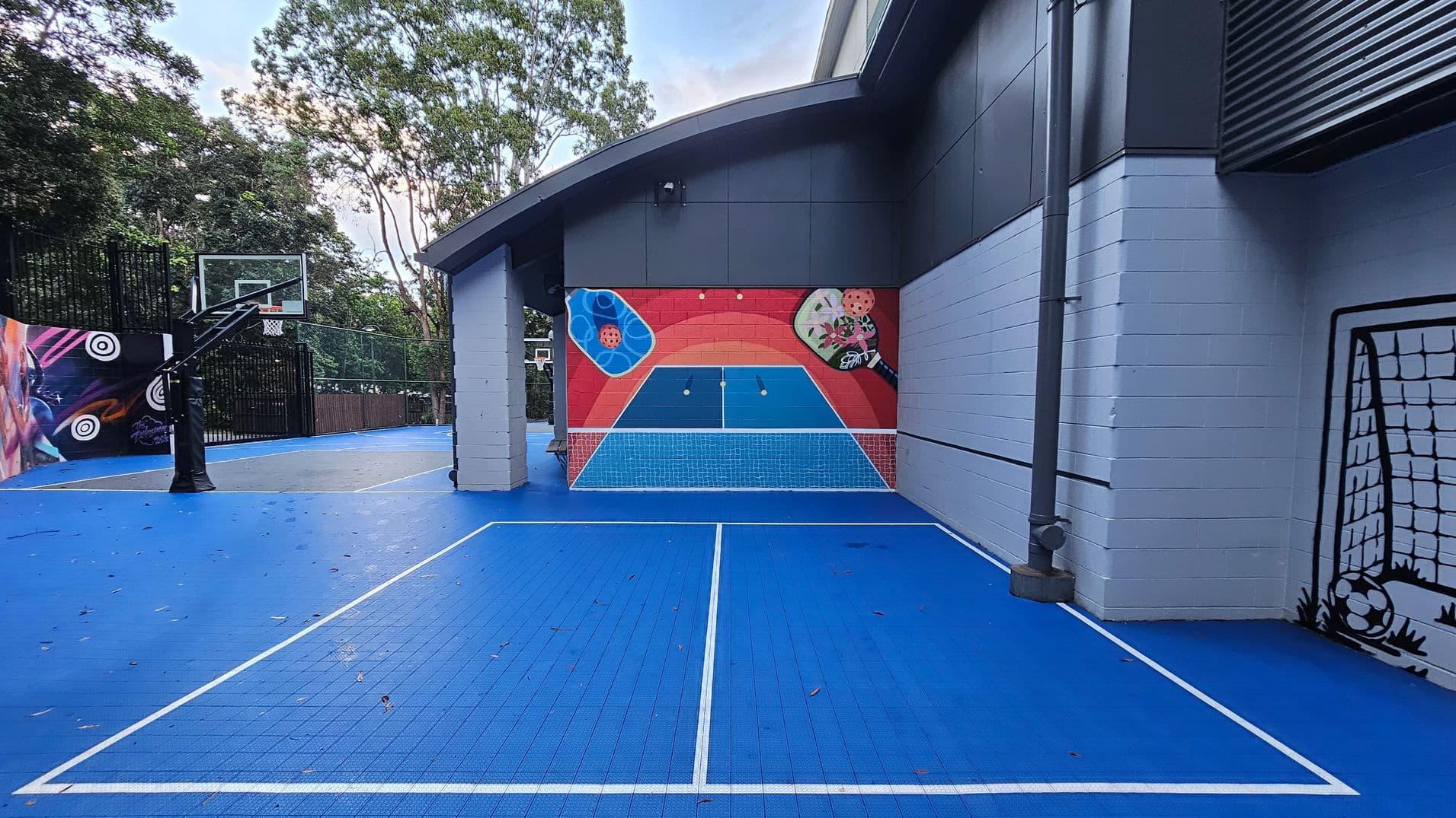 A vibrant sports court with a blue tiled surface and white boundary lines. A basketball hoop stands on the left, adjacent to a colourful mural featuring abstract designs and sports imagery. The court is partially enclosed by a building with grey and white walls. A painted goalpost is visible on the right wall. Trees and a fence line the background, adding a natural boundary to the space.