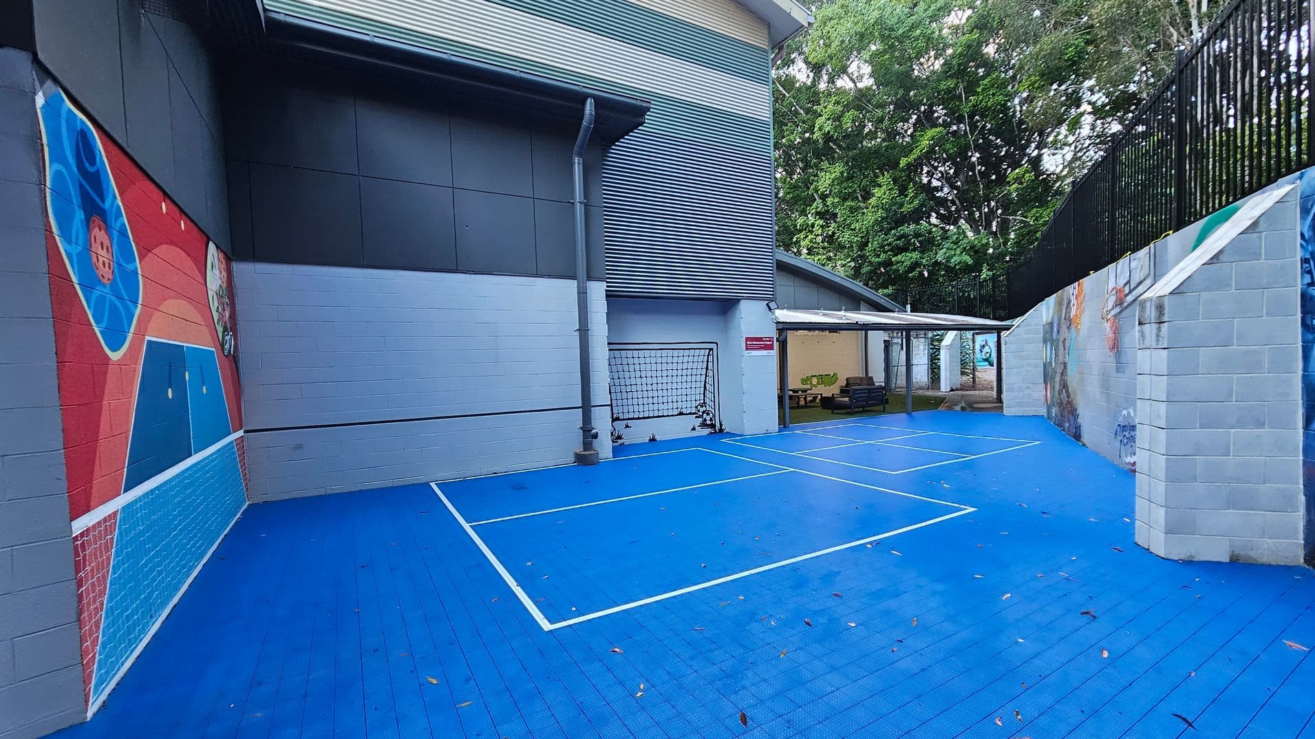 An outdoor sports area with a bright blue court marked with white lines, resembling a small tennis or basketball court. The court is bordered by grey brick walls, one of which features colourful murals. A metal fence runs along the top of one wall. A small goal net is positioned against a wall. To the right, a covered seating area with benches is visible. Trees are seen beyond the fence, adding greenery to the scene.