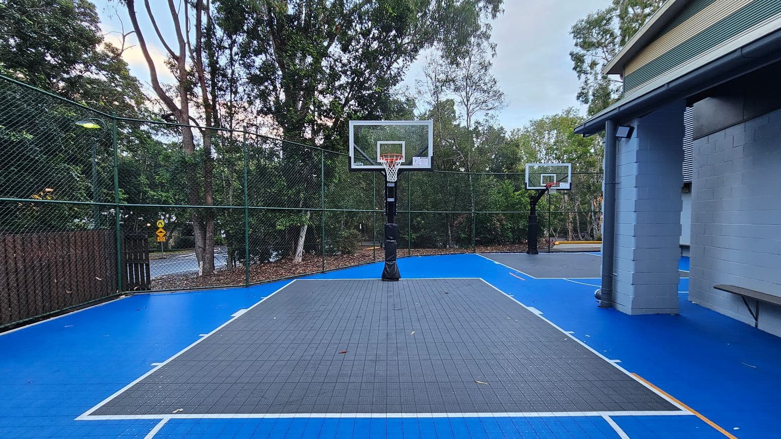 An outdoor basketball court with a vibrant blue surface and a grey key area. Two basketball hoops are visible, one in the foreground and another in the background. The court is enclosed by a green chain-link fence, with tall trees surrounding the area. A building with grey brick walls and a small overhang is on the right. A bench is attached to the building wall. The court is well-lit with overhead lights.