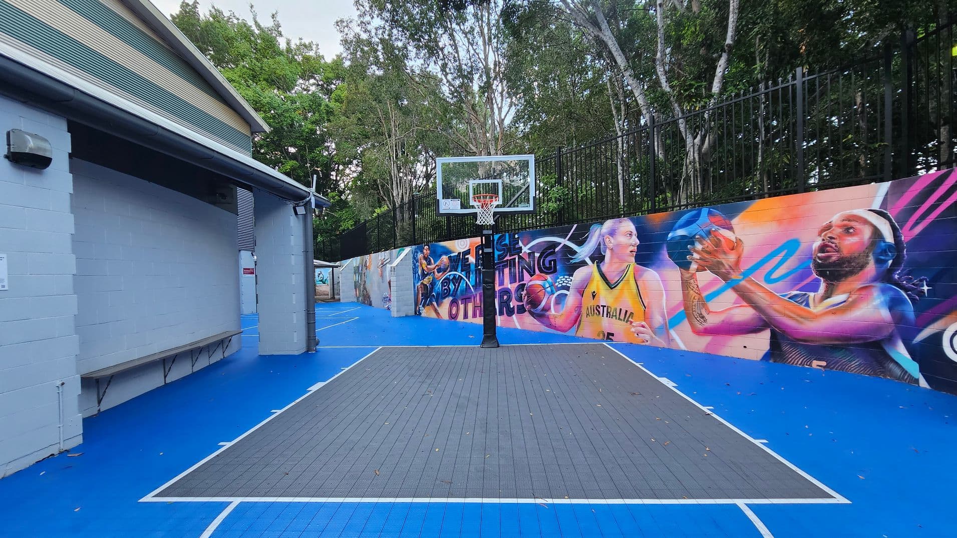 An outdoor basketball court with a vibrant mural on the right wall featuring athletes in action. The court has a blue surface with a grey playing area marked by white lines. A basketball hoop stands centrally at the end of the court. To the left, a grey building with a corrugated metal roof and a bench runs along the wall. Tall trees and a black metal fence form the backdrop, enclosing the space.