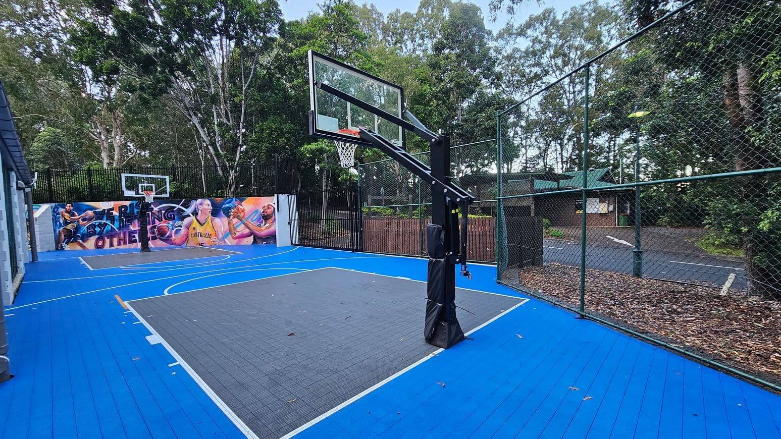 An outdoor basketball court with a vibrant blue surface and grey key area. A basketball hoop with a glass backboard is centrally positioned. The court is enclosed by a green chain-link fence. A colourful mural featuring athletes is painted on the wall at the far end. Trees surround the area, providing a natural backdrop. A small building with a green roof is visible beyond the fence.