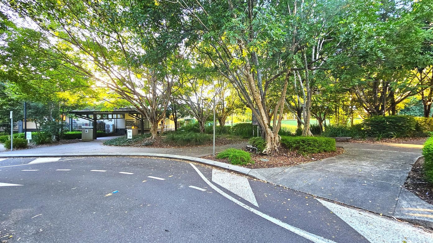 A park-like setting with a paved path curving through the scene, bordered by lush green trees and shrubs. To the left, a small building with a flat roof and large windows is partially visible, surrounded by more greenery. The path is flanked by a low curb and transitions from asphalt to concrete. A metal pole stands near the path, and a bench is nestled among the trees on the right.