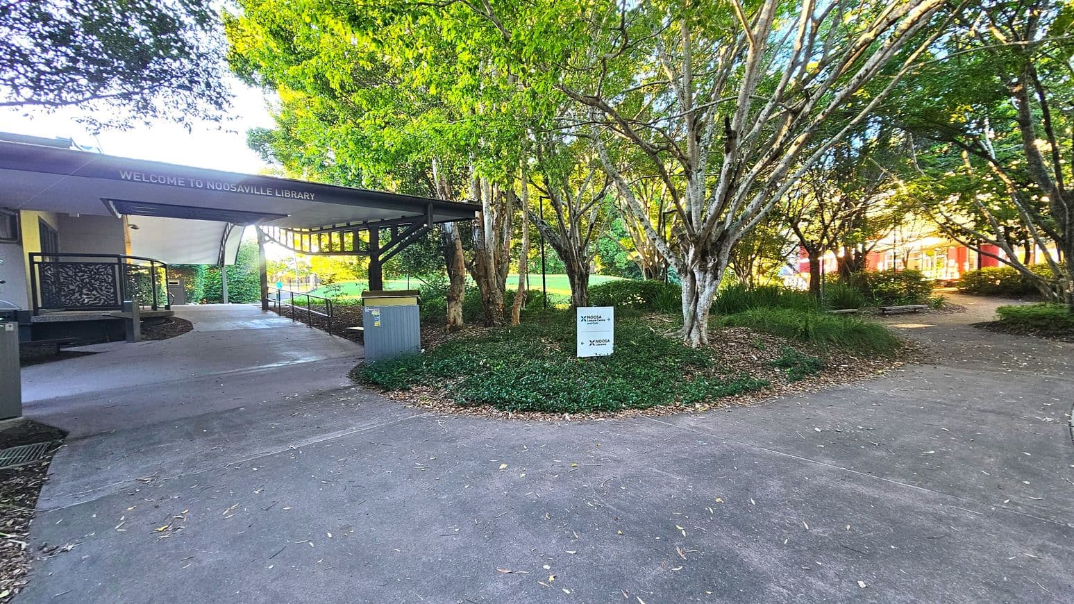A pathway leads to the entrance of Noosaville Library, marked by a sign on a canopy reading 'Welcome to Noosaville Library'. The path is bordered by lush greenery and trees, creating a shaded area. To the right, a small sign with 'Noosa' branding is visible among the foliage. The library building is partially visible on the left, with a modern design featuring metal railings and a covered entrance.