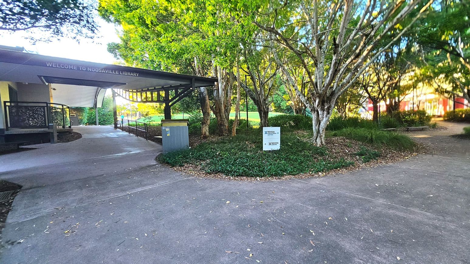 A pathway leads to the entrance of Noosaville Library, marked by a sign above the entrance. The library is partially visible on the left, with a modern design featuring a covered walkway and decorative metal panels. To the right, a cluster of trees and a small sign for Noosa Council are visible. The area is surrounded by greenery, with a mix of concrete and natural ground surfaces.