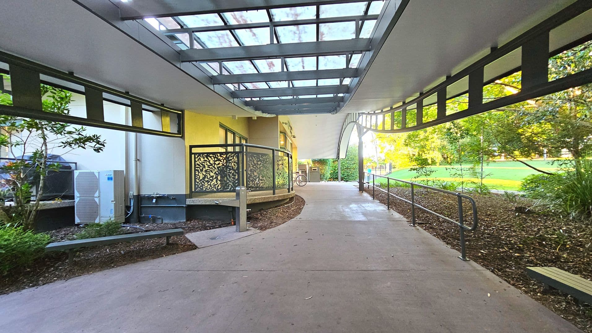 A covered walkway with a transparent roof supported by metal beams, leading to an open area with greenery. On the left, a building with decorative metal railings and a curved path. An air conditioning unit is visible against the wall. On the right, a metal handrail runs along the edge of a mulched garden bed. The path is concrete, transitioning smoothly from the building to the open space.