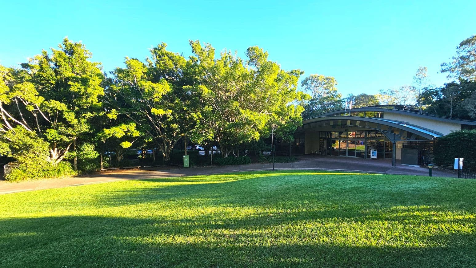 A gently sloping grassy area leads to a modern building with a curved roof and large glass windows. The building is partially obscured by lush green trees on the left. A paved path runs parallel to the building, bordered by a low hedge. A single lamppost stands near the path. The area feels open with a mix of natural and architectural elements.