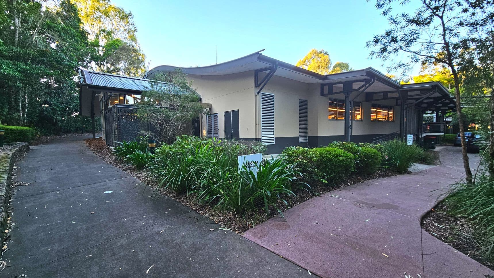 A modern building with a curved roof and large windows is surrounded by lush greenery. Two paths lead to the entrance, one paved with concrete and the other with a reddish surface. The building has beige walls with grey accents and louvred vents. A sign with 'Noosa' is visible among the plants. The area is enclosed by trees and shrubs, creating a natural boundary.