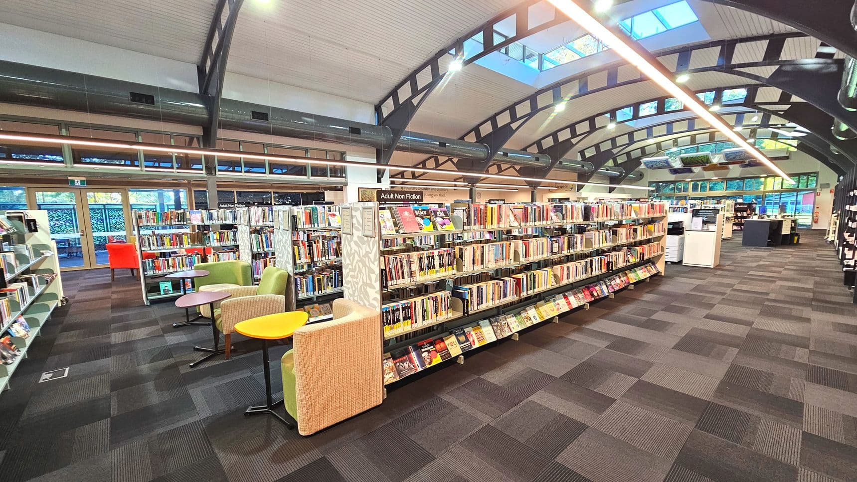 A spacious library interior with high arched ceilings featuring exposed beams and skylights. Rows of bookshelves filled with books are centrally aligned, with a sign indicating 'Adult Non Fiction'. Comfortable seating with colourful chairs and small tables is arranged near the shelves. The floor is covered with patterned carpet tiles. Large windows line the walls, allowing natural light to illuminate the space.