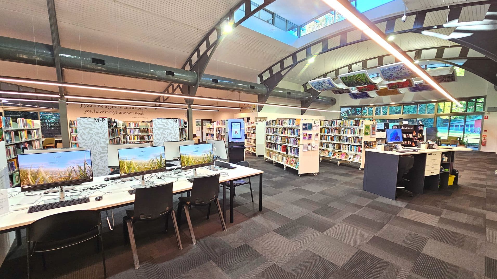 A modern library interior with a high, curved ceiling featuring exposed beams and skylights. In the foreground, a row of computer workstations with large monitors is set on a white desk with black chairs. To the right, a dark-coloured service desk is visible. Bookshelves filled with books line the back wall. The floor is covered with patterned carpet tiles, and large windows on the right allow natural light in.