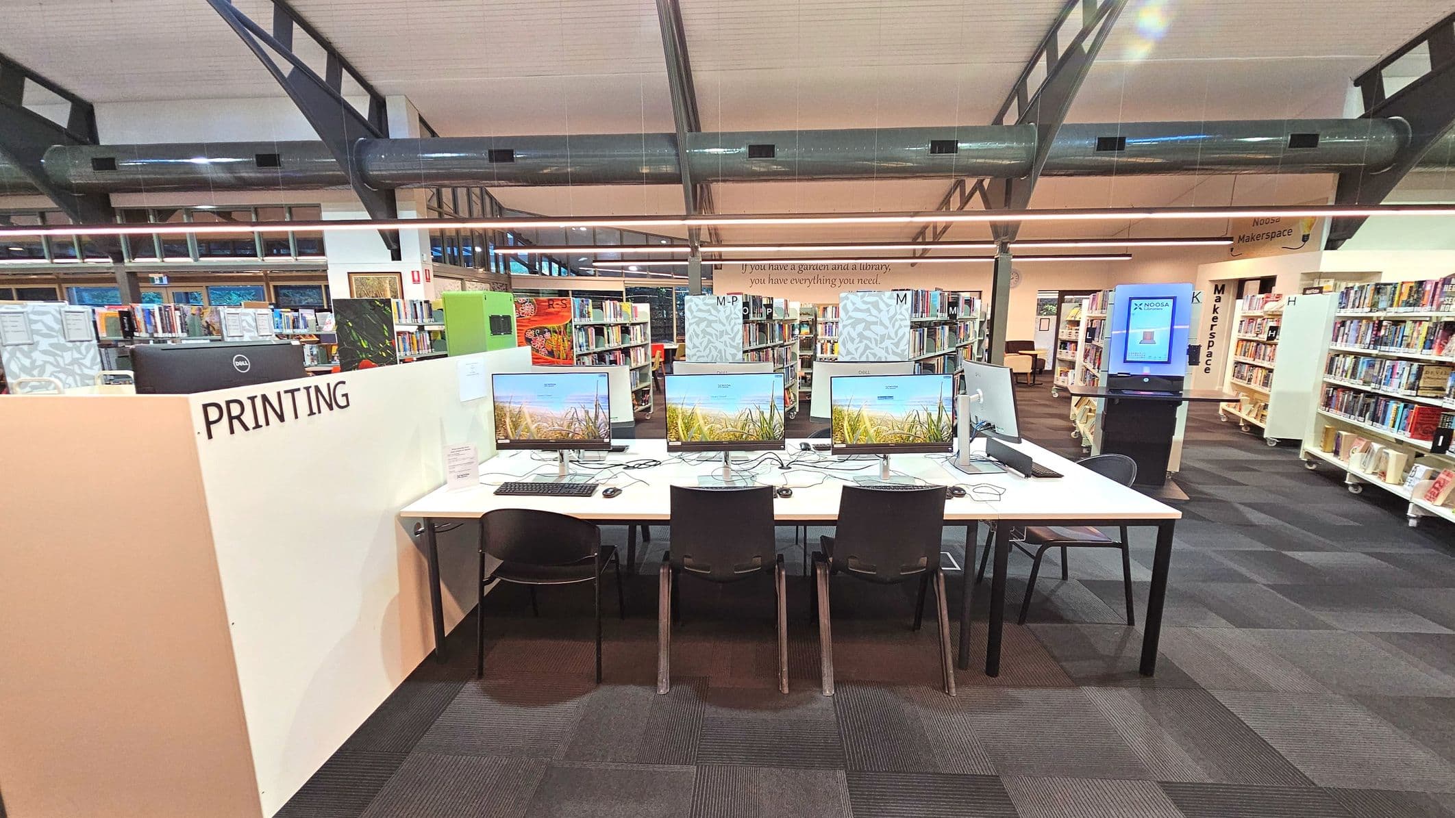 A library interior featuring a row of computers on a white desk with black chairs. A sign reading 'PRINTING' is visible on a partition. Bookshelves filled with books line the background, and a digital kiosk stands to the right. The ceiling has exposed ductwork and large windows. The floor is covered with dark patterned carpet tiles, and a quote is visible on the wall above the bookshelves.