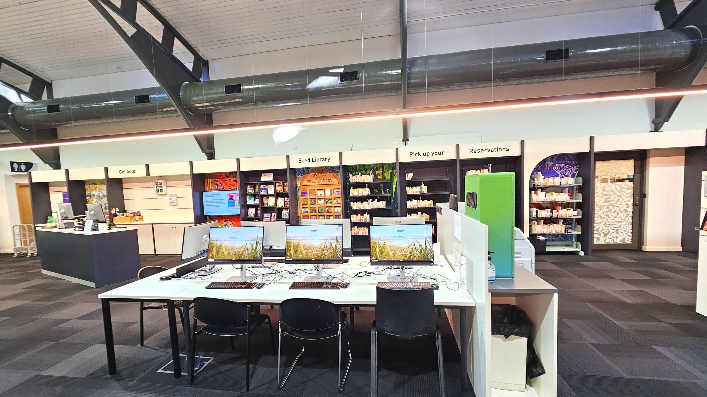 A modern library interior featuring a row of computer stations with monitors and keyboards on a white table. Behind them, shelves labelled 'Seed Library', 'Pick up your', and 'Reservations' are filled with books and seed packets. A 'Get help' desk is visible to the left. The ceiling has exposed ductwork and the floor is covered with dark carpet tiles. A sign for restrooms is seen on the left wall.