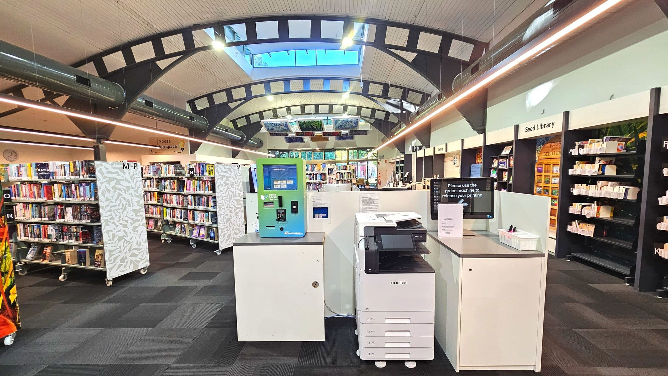 A modern library interior with a high, curved ceiling featuring exposed beams and a skylight. In the foreground, a green self-service machine and a white printer are on a counter. Bookshelves filled with books are arranged in rows, with signage indicating sections like 'M-P'. The floor is covered with dark carpet tiles. To the right, a section labelled 'Seed Library' is visible, with shelves displaying various items.