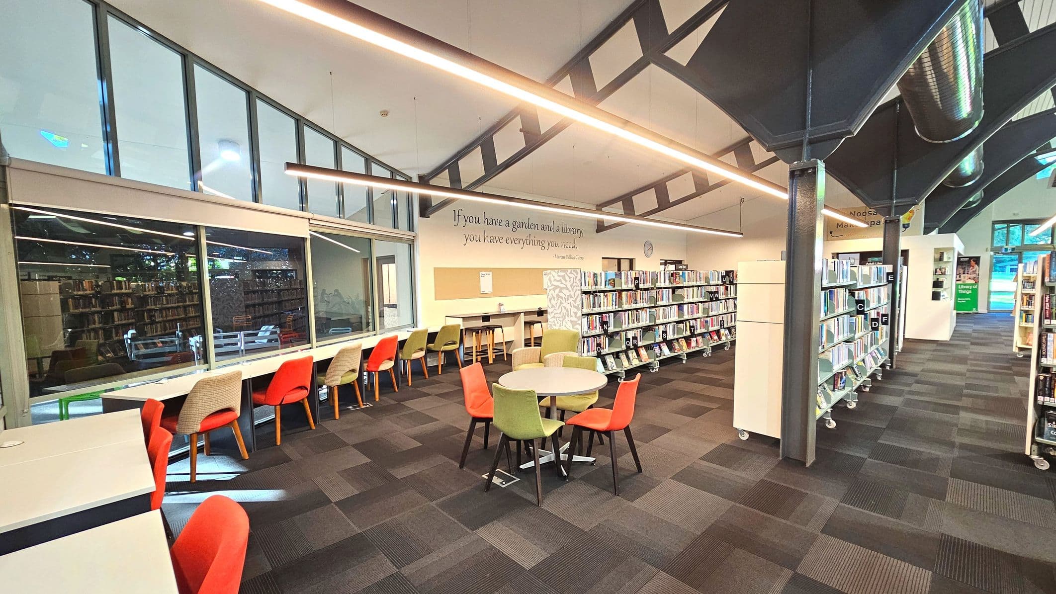 A modern library interior with high ceilings and exposed beams. On the left, a row of desks with colourful chairs faces large windows. A quote is displayed on the wall above. In the centre, a round table with mixed chairs sits on a patterned carpet. To the right, bookshelves on wheels are neatly arranged. The space is well-lit with linear ceiling lights and features a mix of glass and metal elements.