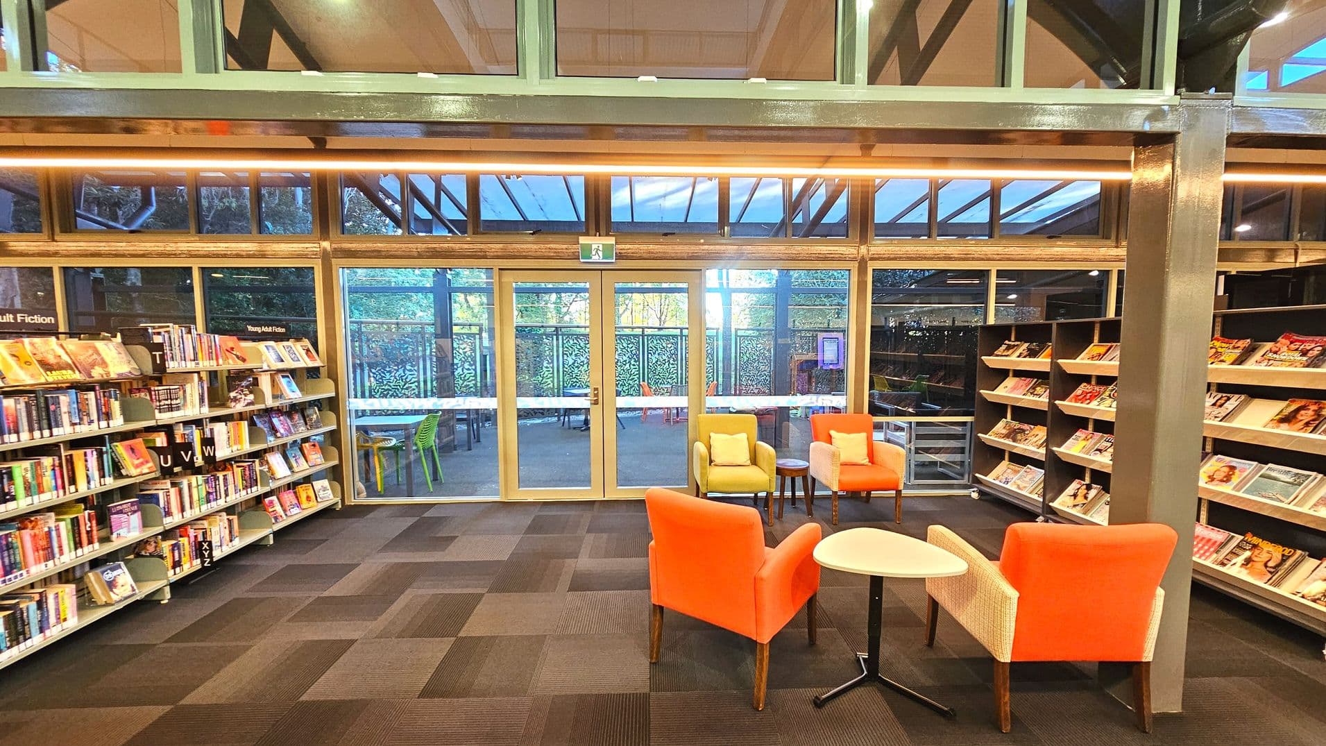 A library interior with bookshelves on the left and right filled with books and magazines. In the centre, there are two orange and one yellow armchairs around small round tables. The floor is covered with patterned carpet tiles. Large glass doors and windows at the back reveal an outdoor area with green chairs and tables. The ceiling features exposed beams and lighting. A sign above the door indicates an exit.