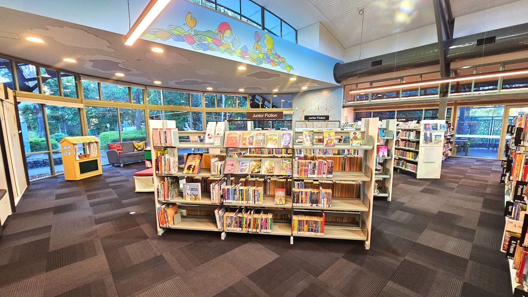 A library interior featuring a section labelled 'Junior Fiction' with several white bookshelves filled with colourful children's books. The floor is covered with dark patterned carpet tiles. Large windows with wooden frames line the curved wall, offering a view of greenery outside. A small wooden playhouse and a grey sofa are visible near the windows. Overhead, a decorative panel with cartoon characters is mounted on the ceiling.