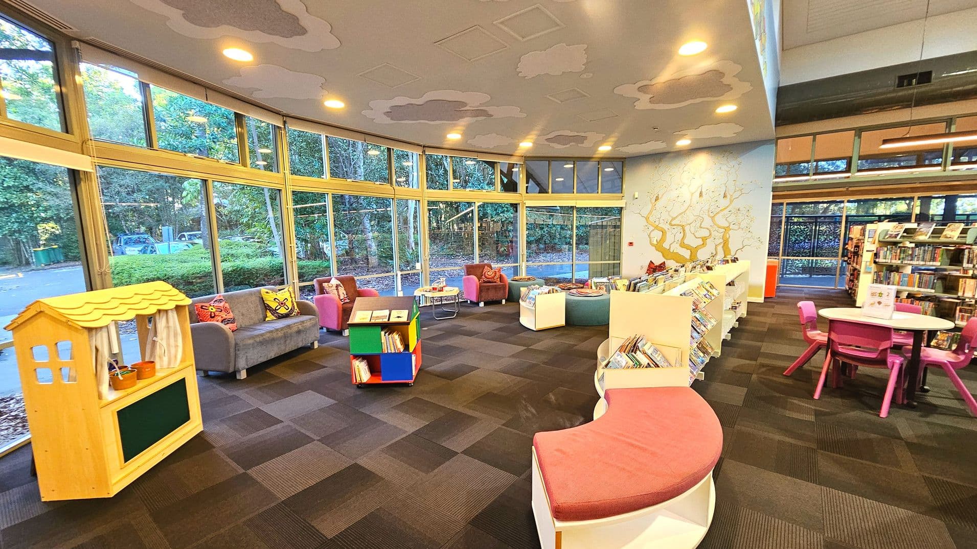 A spacious library area with large floor-to-ceiling windows curving along one side, allowing natural light to flood in. The room features a variety of seating options including colourful armchairs and a grey sofa with decorative cushions. A small wooden puppet theatre stands near the windows. Books are displayed on low, white shelves and a round table with pink chairs is visible. The ceiling has cloud-like patterns, and a mural decorates one wall.