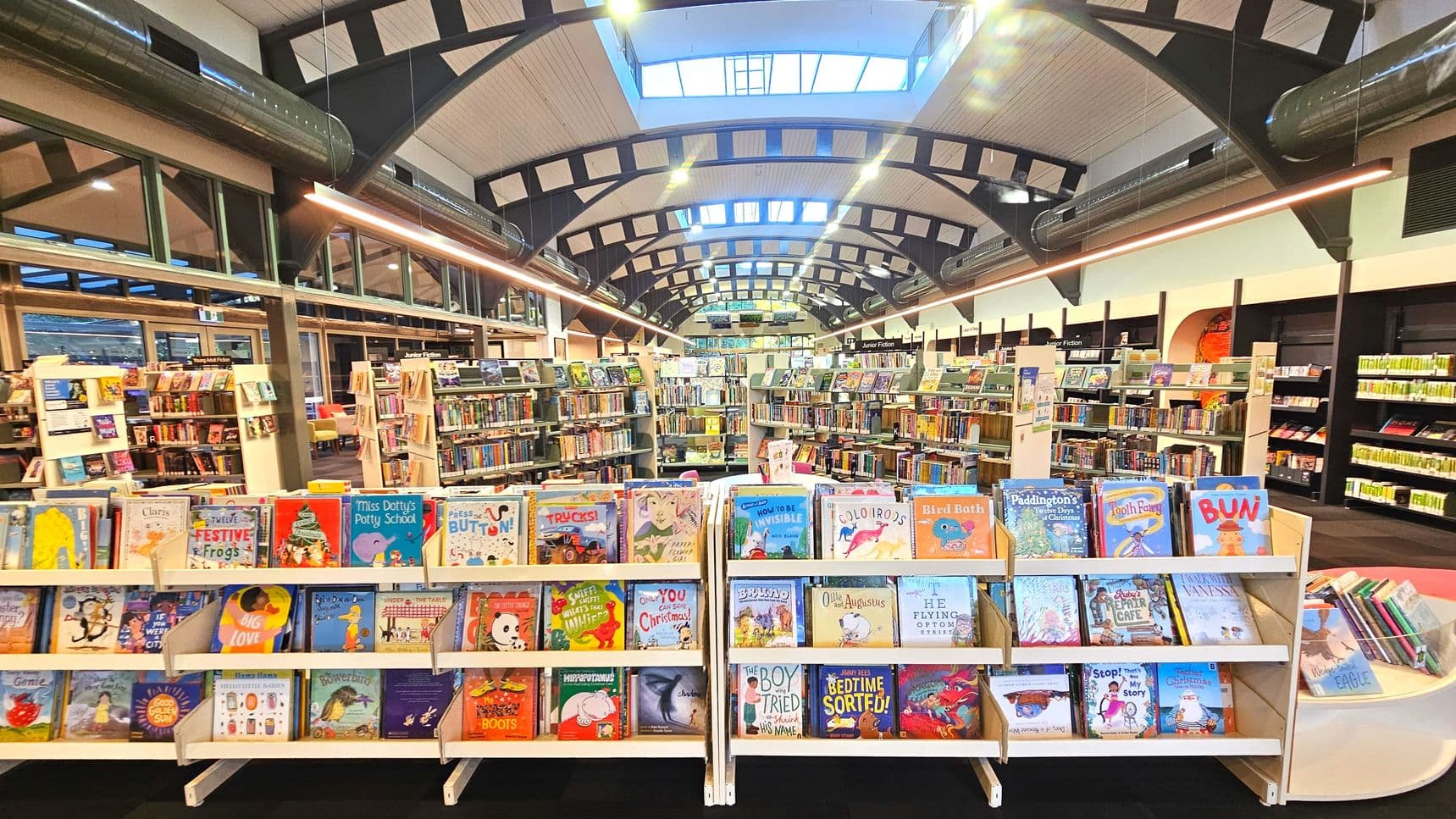 A spacious library interior with a high arched ceiling featuring exposed beams and skylights. In the foreground, there are several white bookstands displaying colourful children's books. Behind them, rows of bookshelves are filled with various books. The floor is carpeted, and the space feels open with large windows on the left. Overhead, long cylindrical ducts run parallel to the ceiling beams.