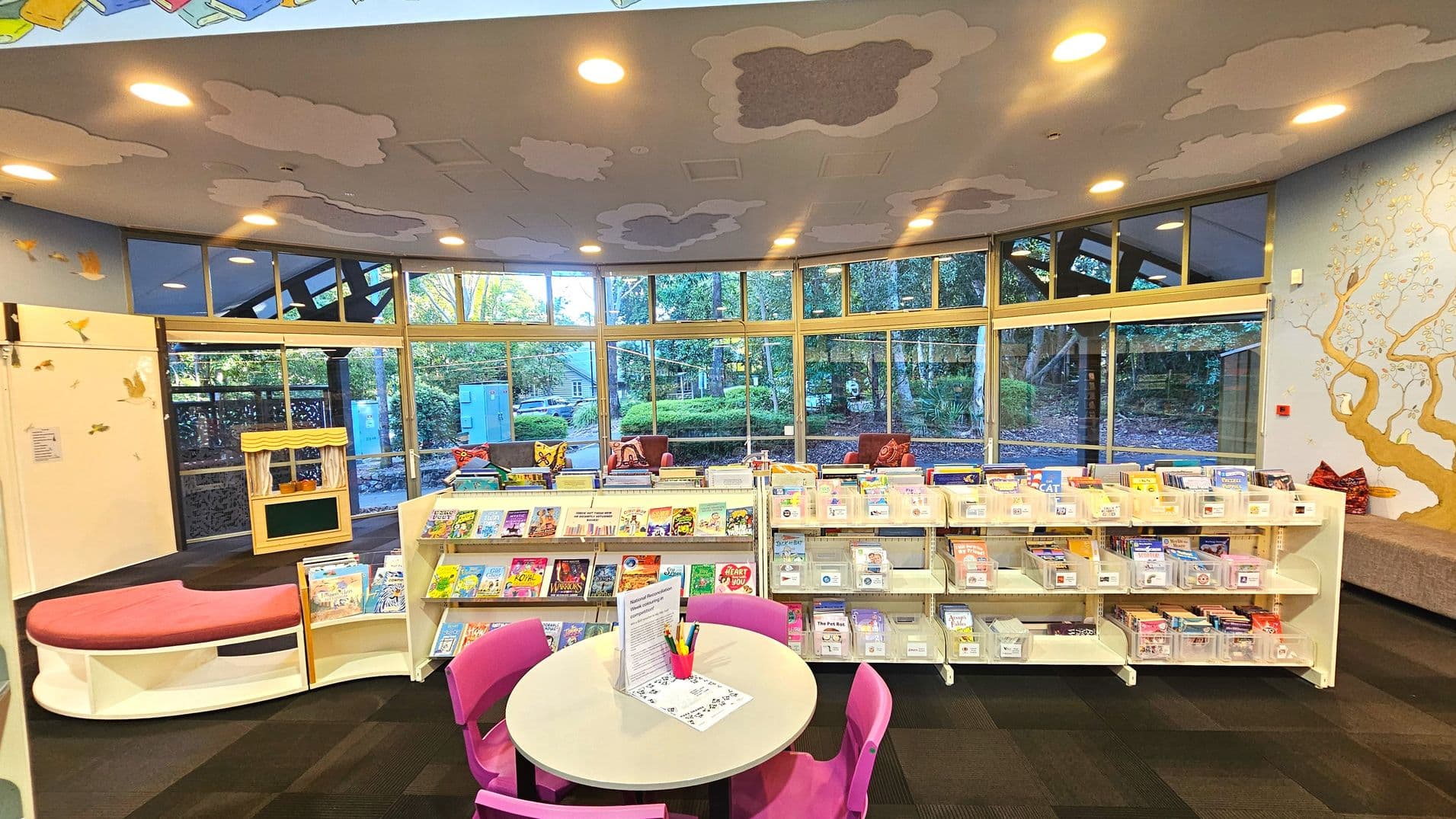 A children's library area with large windows curving along the back wall, offering a view of greenery outside. Inside, there are low white bookshelves filled with colourful children's books. A round table with pink chairs is in the foreground. The ceiling features cloud-like decorations, and a mural of a tree adorns one wall. The floor is carpeted in dark squares, and a small puppet theatre is visible to the left.