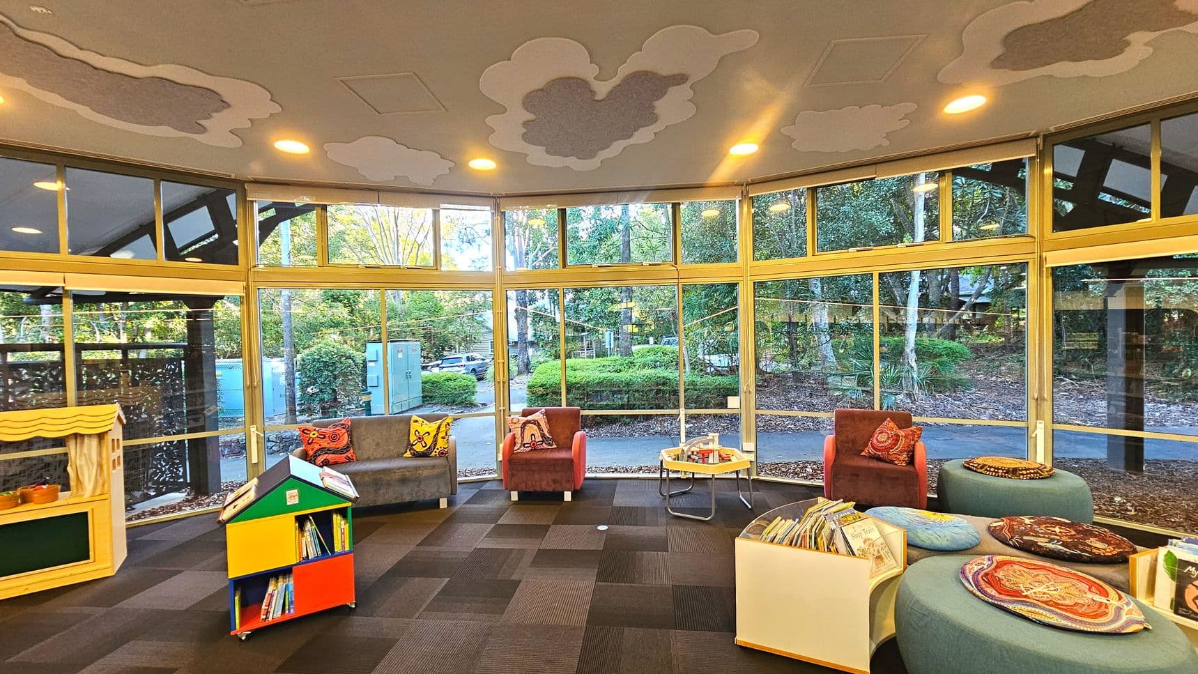 A circular room with large floor-to-ceiling windows offering a view of lush greenery outside. The interior features a variety of colourful furniture, including a small puppet theatre, a multi-coloured book display, and several cushioned chairs and ottomans. The ceiling is decorated with cloud patterns, and the floor is covered with dark carpet tiles. The space is well-lit with recessed lighting.