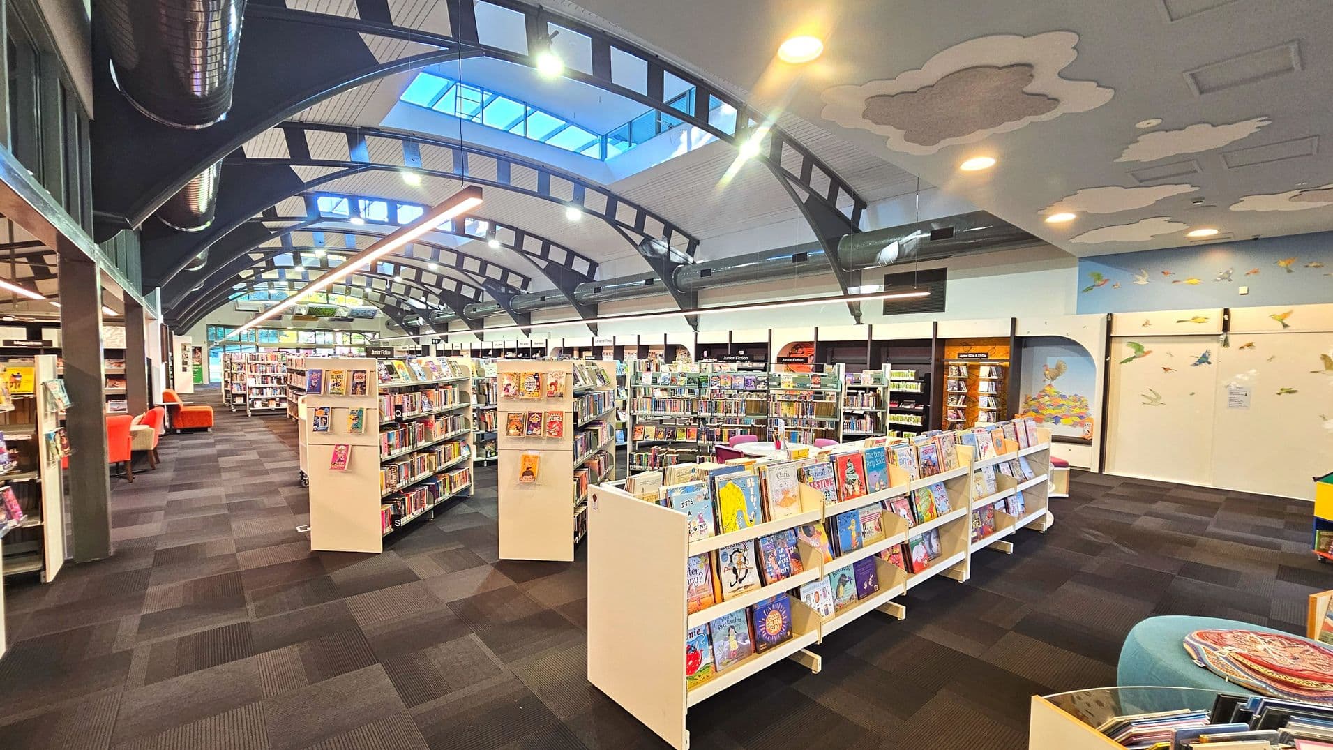 A spacious library interior with a high, arched ceiling featuring skylights and exposed beams. Rows of bookshelves filled with colourful books are neatly arranged on a dark carpeted floor. Bright orange chairs are visible to the left, and the walls are adorned with playful murals of birds and clouds. The space feels open and inviting, with ample lighting from overhead fixtures and natural light from the skylights.