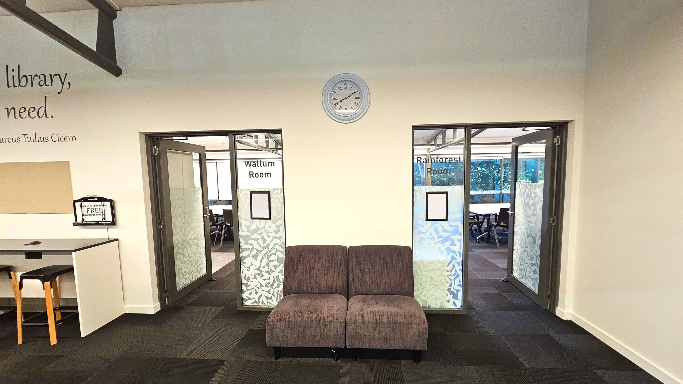 A hallway with two glass doors leading to the Wallum Room and Rainforest Room, each with frosted leaf patterns. A brown two-seater sofa is centred between the doors. Above, a round wall clock is mounted. To the left, a counter with a high stool and a small sign. The carpet is dark with a subtle pattern, and the walls are light-coloured with a quote partially visible.