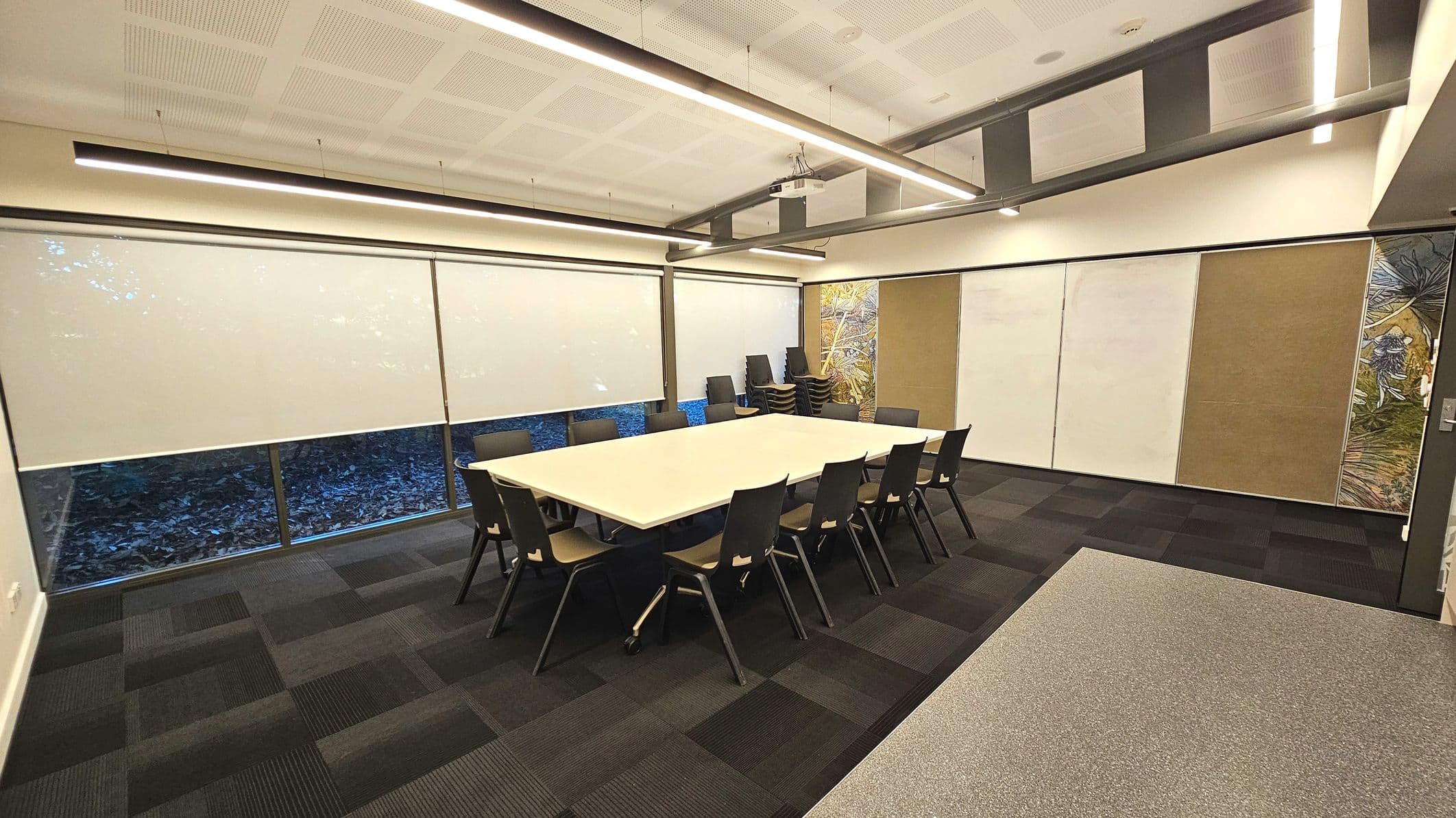 A modern conference room with a large rectangular table surrounded by black chairs. The room features floor-to-ceiling windows with white roller blinds partially drawn. The walls are adorned with panels, some with artwork. The ceiling has recessed lighting and a projector mounted above the table. The floor is covered with dark patterned carpet tiles, transitioning to a lighter grey carpet near the entrance.