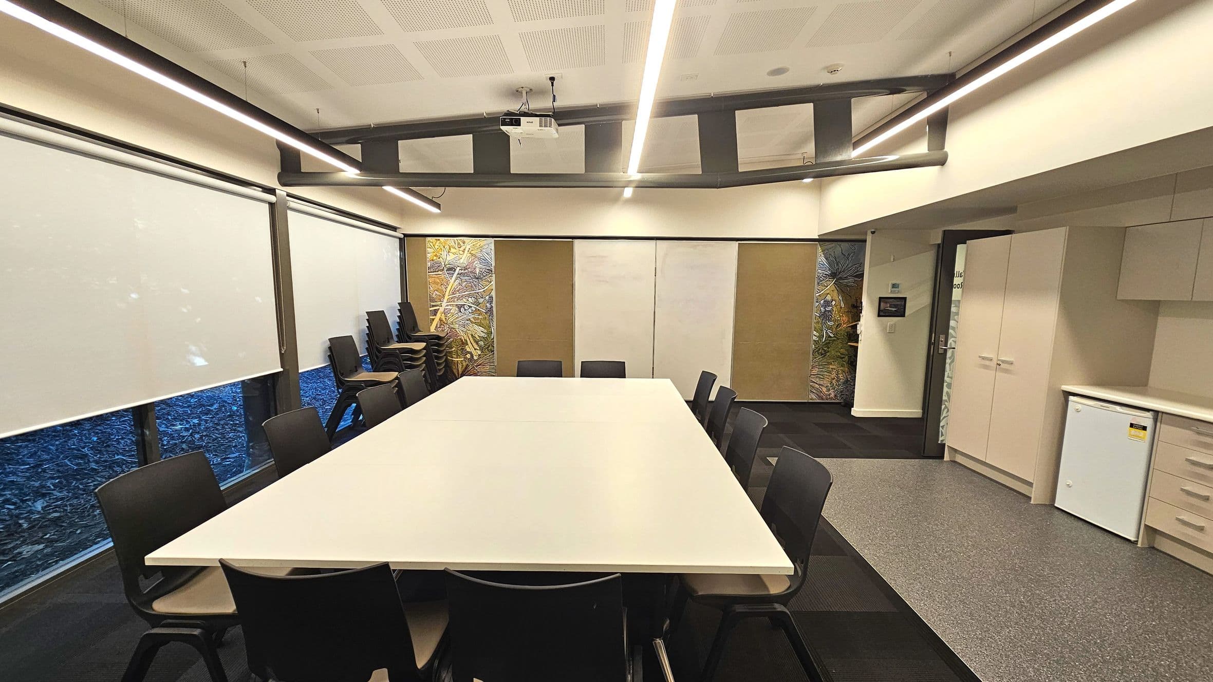 A modern meeting room with a large rectangular table surrounded by black chairs. The room features a ceiling-mounted projector and fluorescent lighting. The walls have decorative panels with nature motifs and a section of white cabinets with a small fridge. The floor transitions from dark carpet to light grey vinyl near the cabinets. Large windows with roller blinds line one side, and a stack of extra chairs is visible.