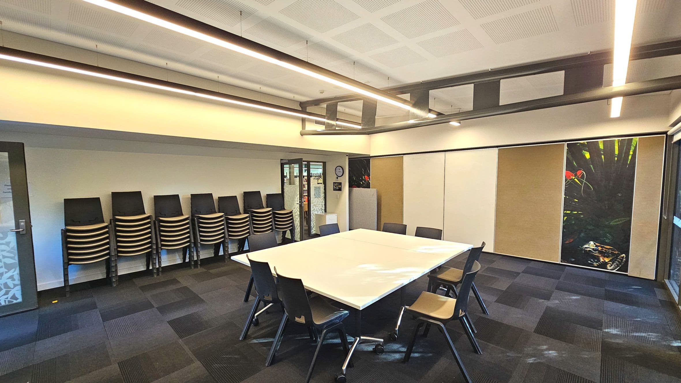 A modern meeting room with a large square table at the centre surrounded by eight chairs. The floor is covered with dark, patterned carpet tiles. On the left, stacks of chairs are neatly arranged against the wall. A glass door with frosted patterns is visible, and a clock is mounted above it. The right wall features a large, vibrant nature-themed mural. The ceiling has recessed lighting and exposed ductwork.