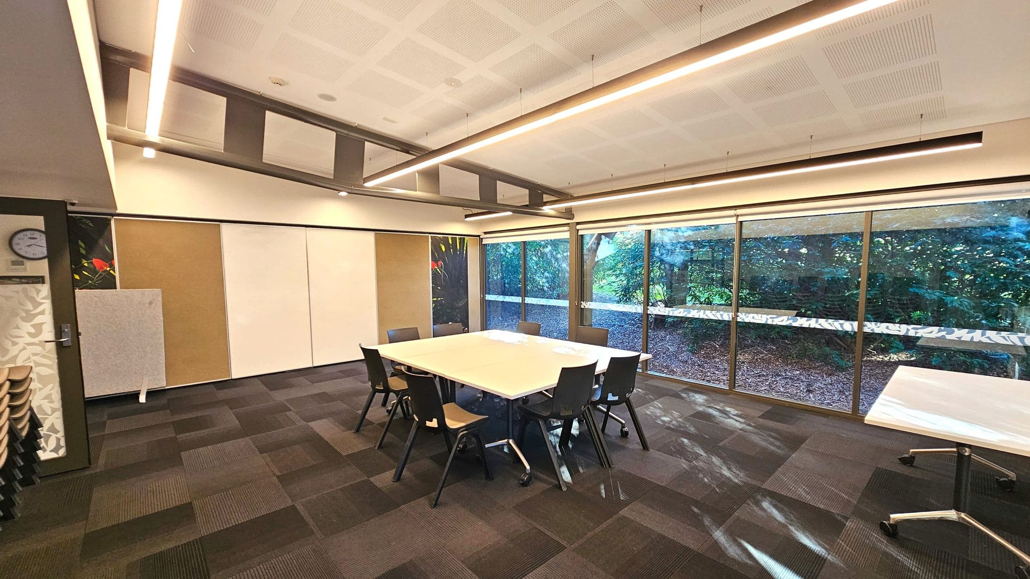 A modern meeting room with a large square table surrounded by eight black chairs on a carpeted floor with a checkered pattern. The room features a wall of floor-to-ceiling windows offering a view of dense greenery outside. The ceiling has recessed lighting and a grid pattern. A clock and a decorative panel are mounted on the left wall, and a stack of chairs is visible in the corner.