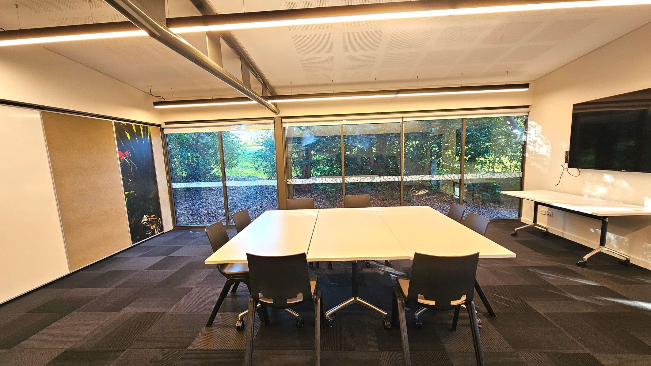 A modern meeting room with a large rectangular table surrounded by eight black chairs on a dark carpeted floor. The room features a wall of floor-to-ceiling windows with views of greenery outside. A large flat-screen TV is mounted on the right wall above a small white table. The ceiling has linear LED lights and exposed ductwork. A colourful abstract artwork is displayed on the left wall.
