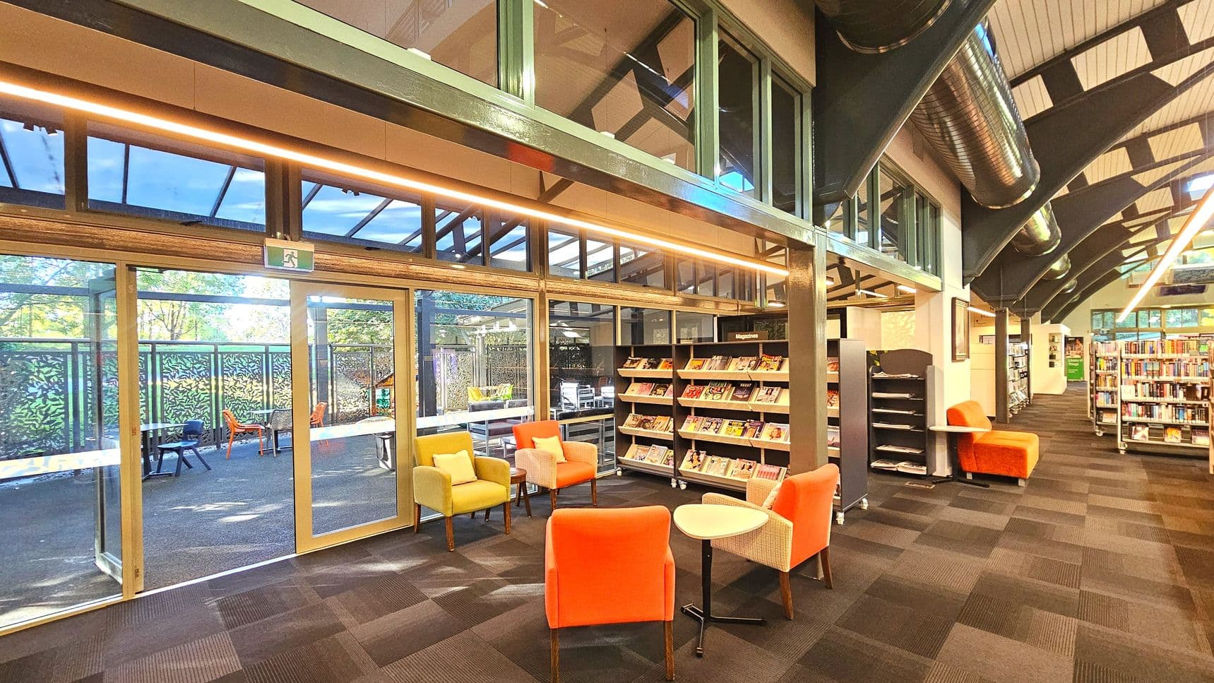 The interior of a modern library features a seating area with colourful armchairs and small tables on a carpeted floor. To the left, large glass doors lead to an outdoor patio with tables and chairs. Bookshelves filled with magazines and books line the walls. Overhead, exposed ductwork and beams are visible, and the ceiling is lined with panels. The space is well-lit with natural light streaming through the windows.