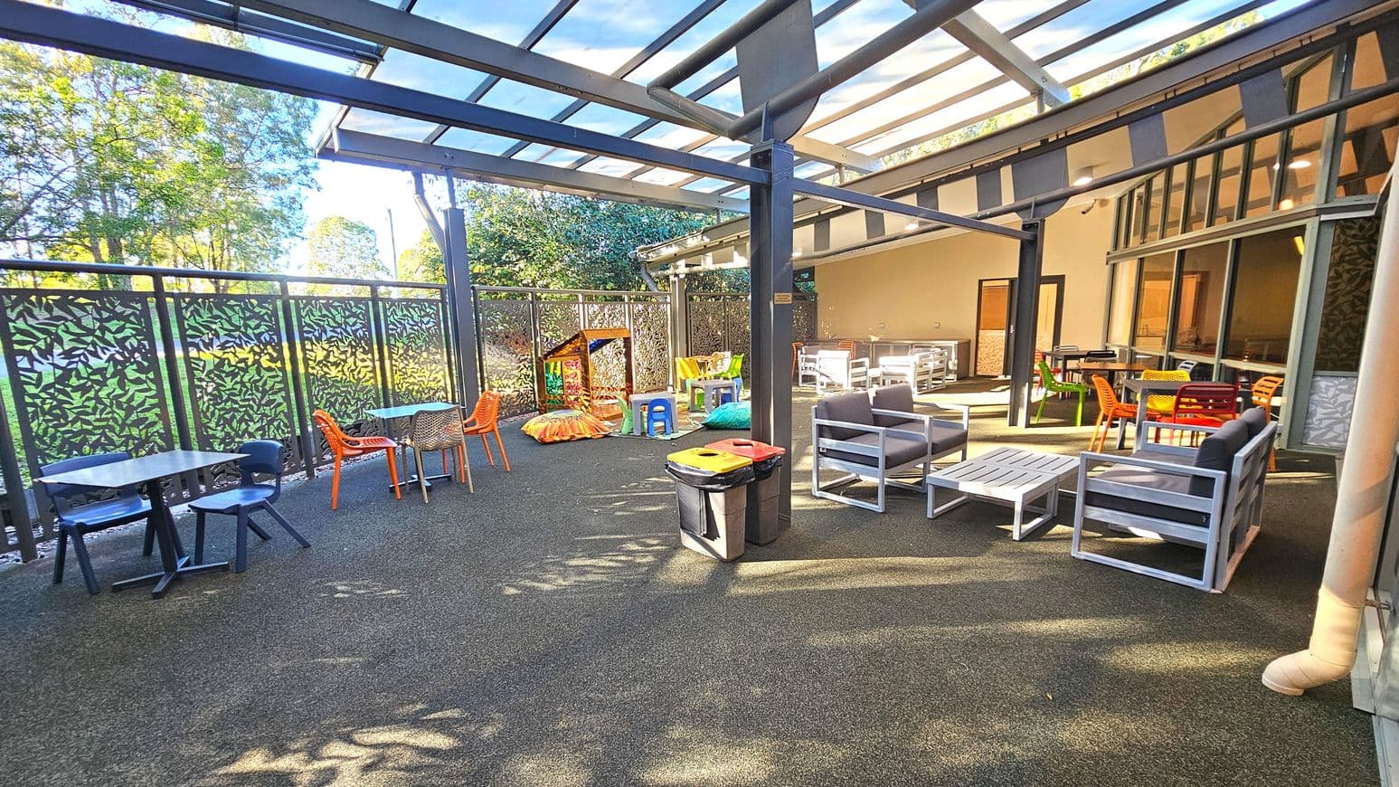 An outdoor seating area with a transparent roof supported by metal beams. The space features various seating options, including grey cushioned chairs with white frames, colourful plastic chairs, and small tables. A decorative metal fence with leaf patterns encloses the area. The floor is a mix of textured surfaces, and there are bins for waste disposal. A children's play area with cushions and a small playhouse is visible.