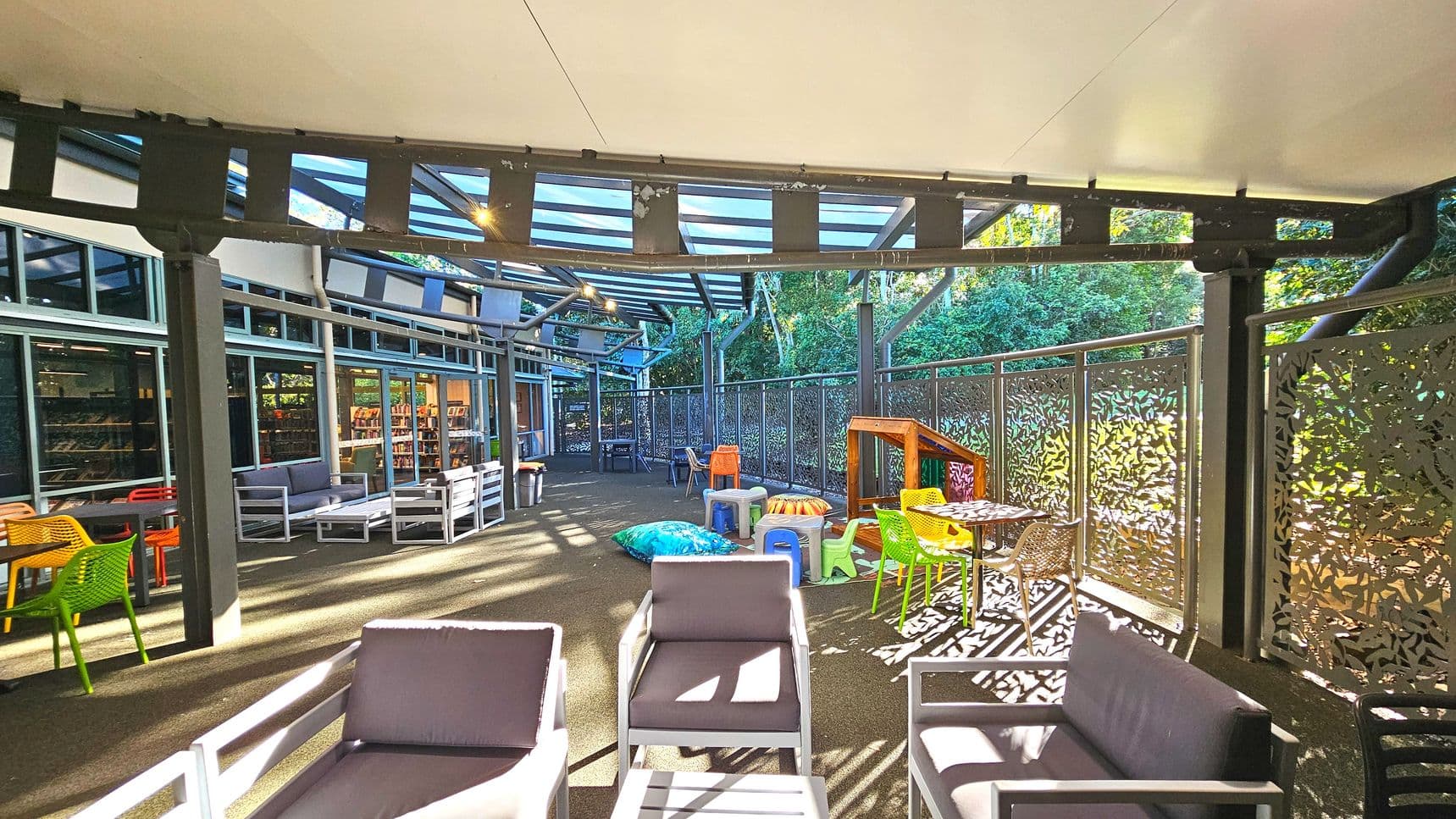 An outdoor seating area with a mix of grey cushioned chairs and colourful plastic chairs and tables. The space is partially enclosed by decorative metal panels with leaf patterns. A transparent roof supported by metal beams provides shelter. The ground is covered with dark paving, and there are children's play items like a small slide and bean bags. Large windows on one side reveal an indoor area with bookshelves.