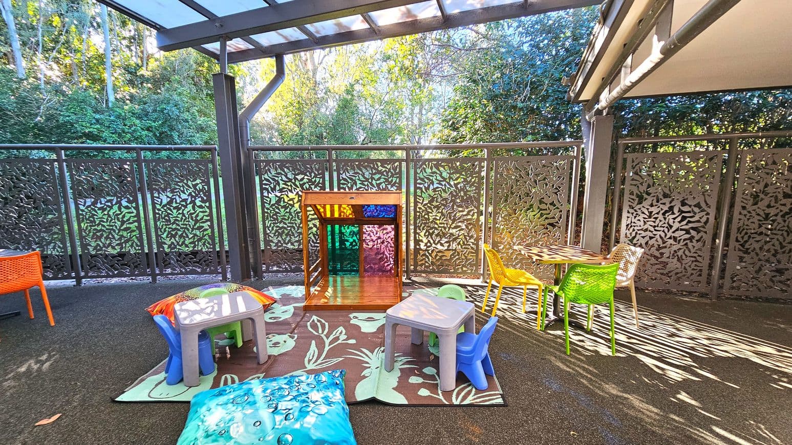 An outdoor play area with a transparent roof and decorative metal fencing featuring leaf patterns. The space includes a small wooden playhouse with colourful panels, surrounded by plastic tables and chairs in bright colours like orange, green, and blue. A patterned rug with plant designs covers part of the rubberised floor. The area is bordered by lush greenery, providing a natural backdrop.