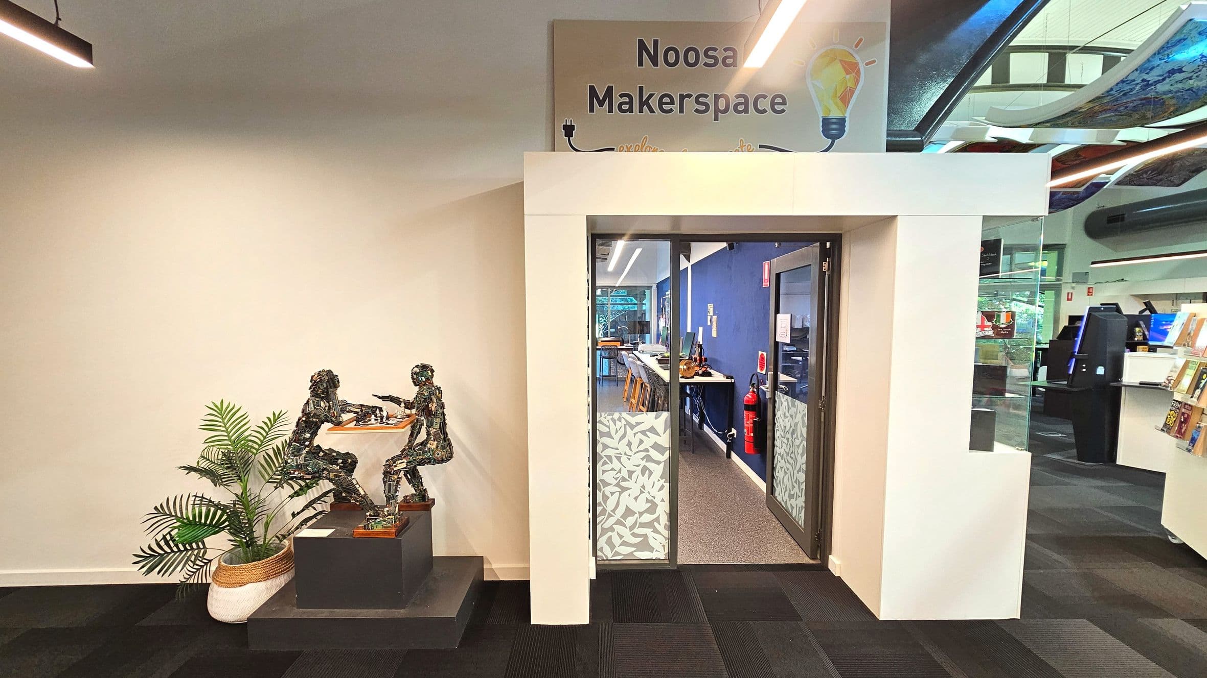 The image shows the entrance to the Noosa Makerspace, featuring a sign above the doorway. The entrance is framed by white walls and glass doors with a leaf pattern. Inside, a blue wall is visible with tables and stools. To the left, a sculpture of two figures playing chess is displayed on a pedestal next to a potted plant. The floor transitions from dark carpet to a lighter speckled surface inside.